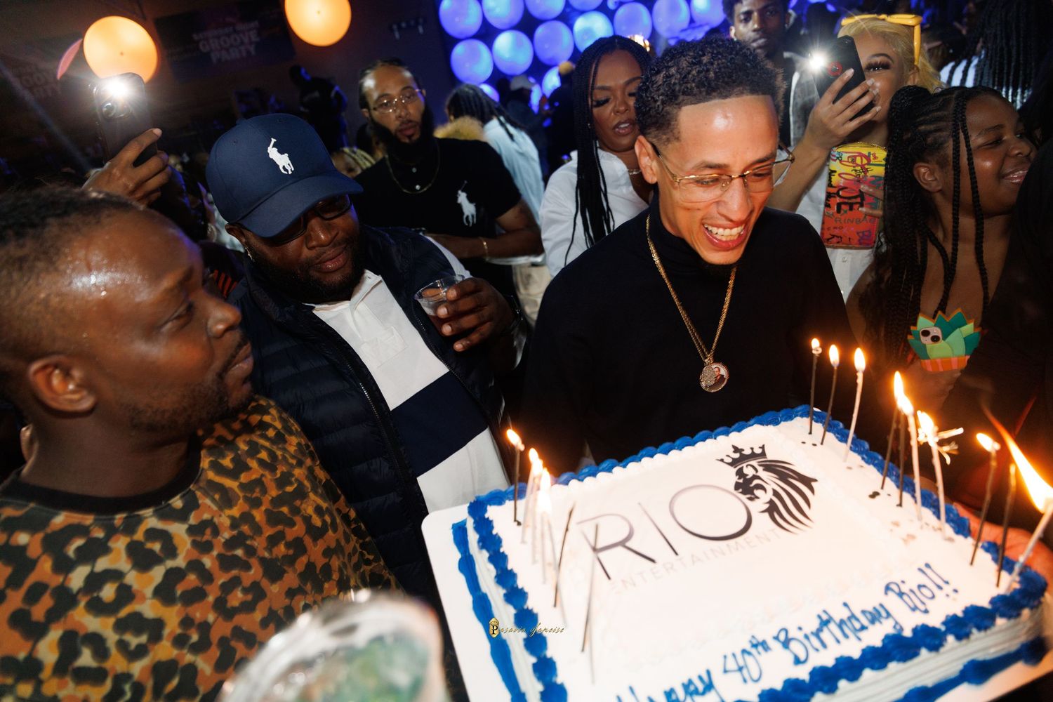 Group of people celebrating DMario Grey birthday party with a large decorated cake with lit candles at a festive indoor event.