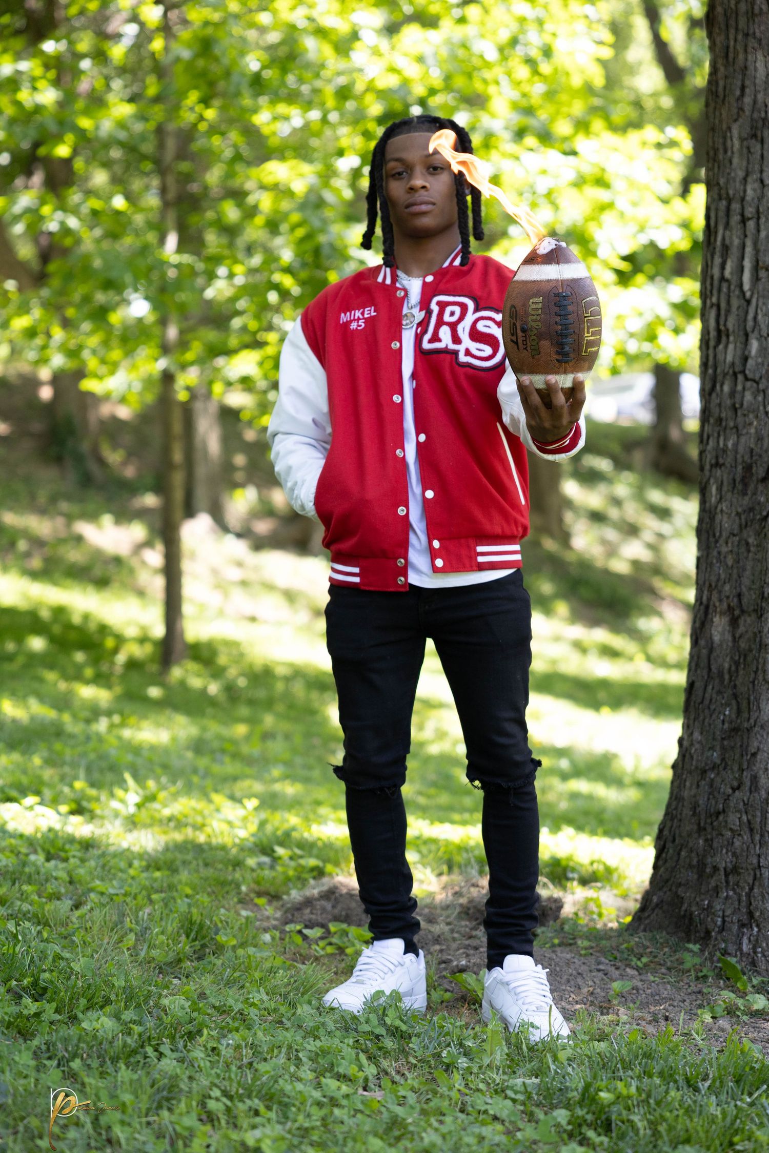 Young man in red varsity jacket holding football on fire outdoors near tree in green park setting.
