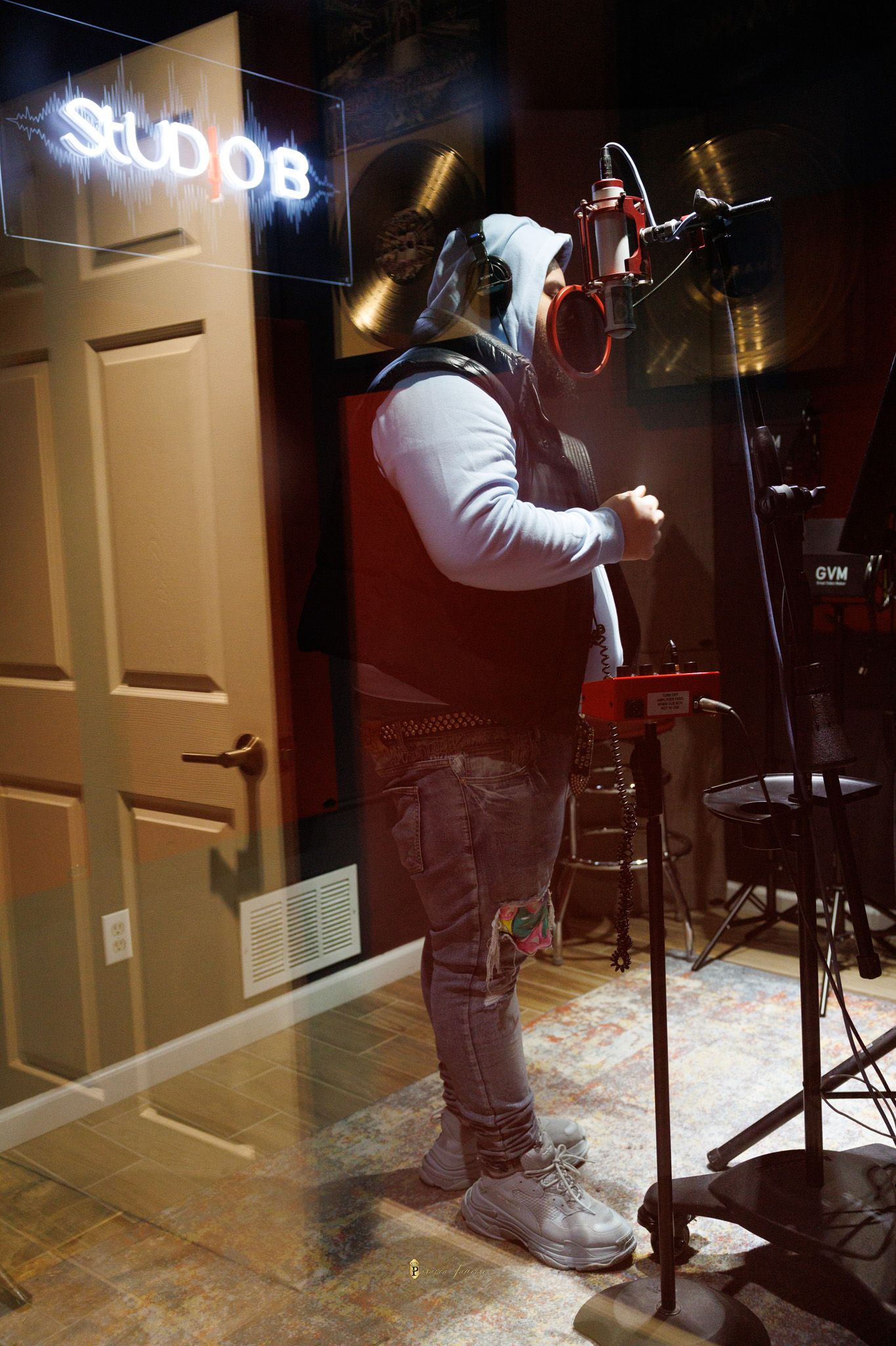 Artist recording in a professional music studio, standing at a red microphone on a stand with neon Studio sign in background.