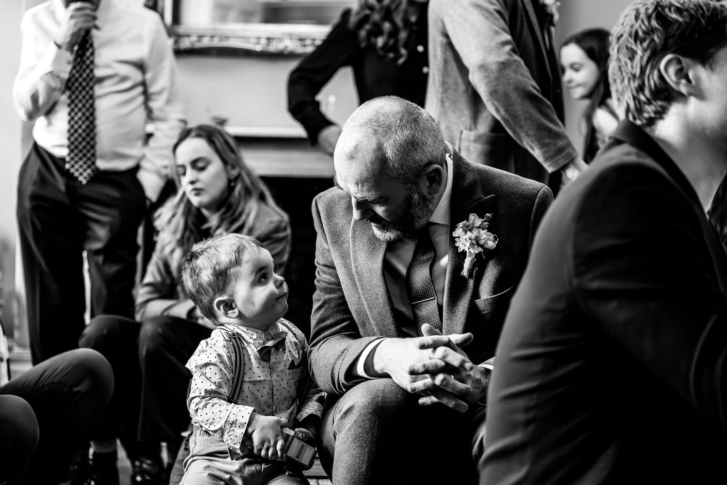 Black and white wedding photo of a bald man in a suit interacting warmly with a toddler at a wedding reception.