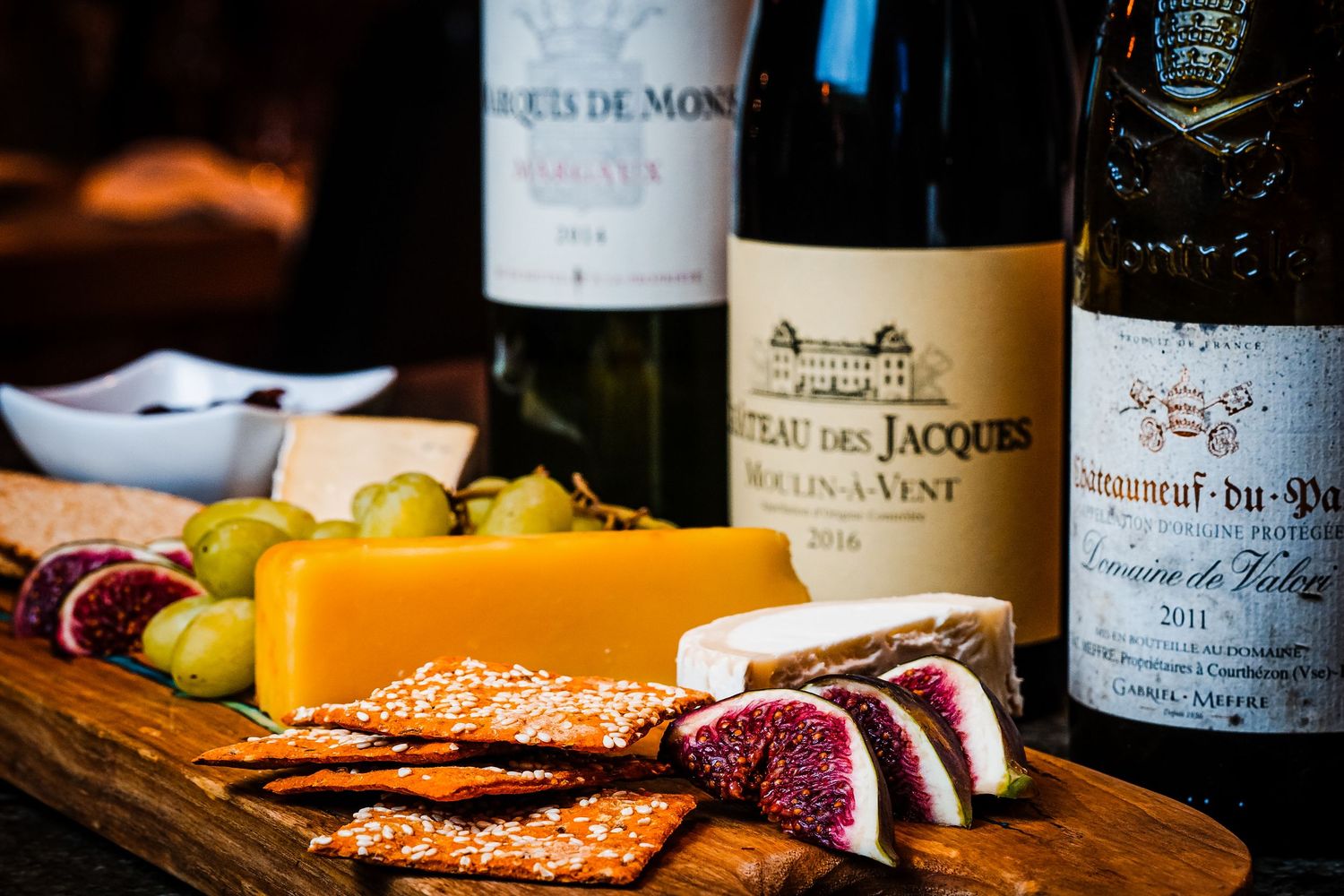 A rustic wooden board with cheese, figs, crackers, grapes, and three bottles of French red wine.