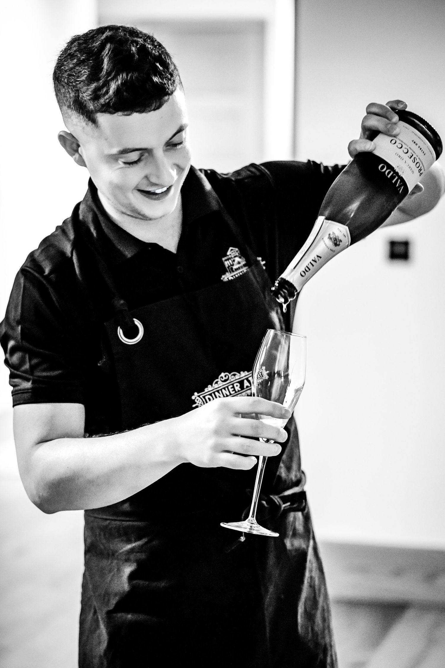 Smiling bartender in black apron pouring prosecco into a champagne flute, black and white photo.