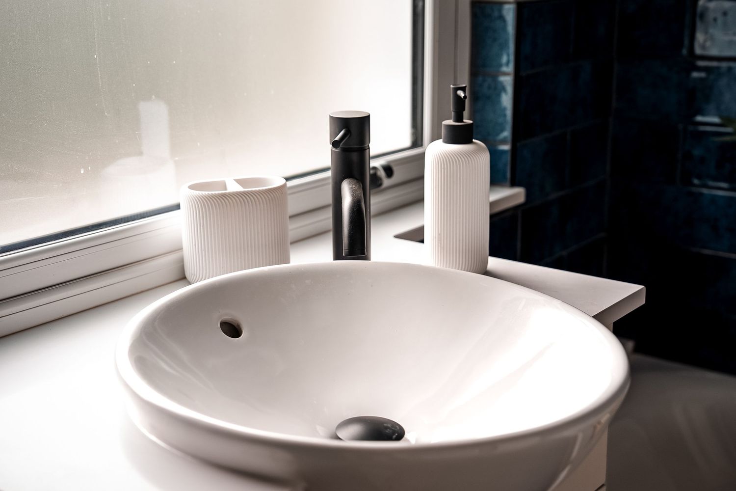 Modern white round vessel sink with black faucet, soap dispenser, and candle on a white countertop near a window.