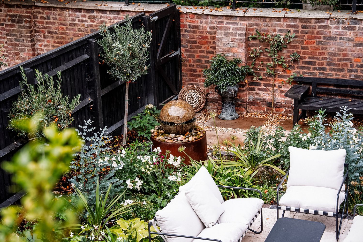 Lush courtyard garden with white outdoor furniture, fountain, and greenery against brick wall and black fence.