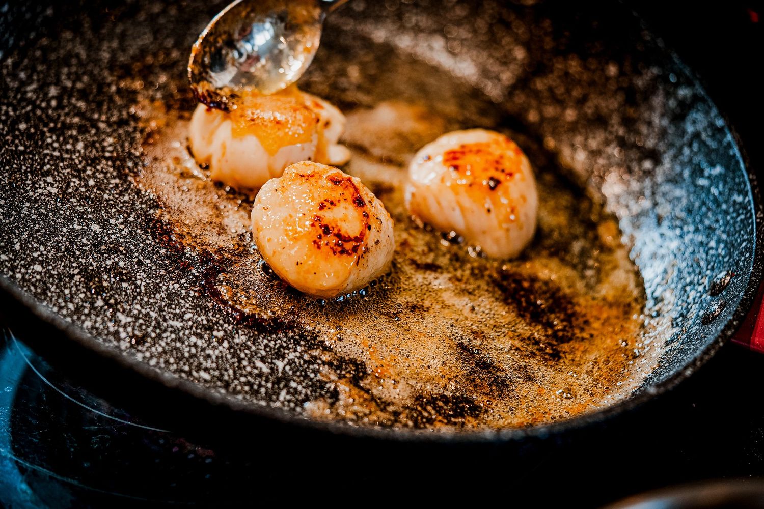 Three golden-seared scallops cooking in a dark skillet with bubbling butter and caramelized fond.