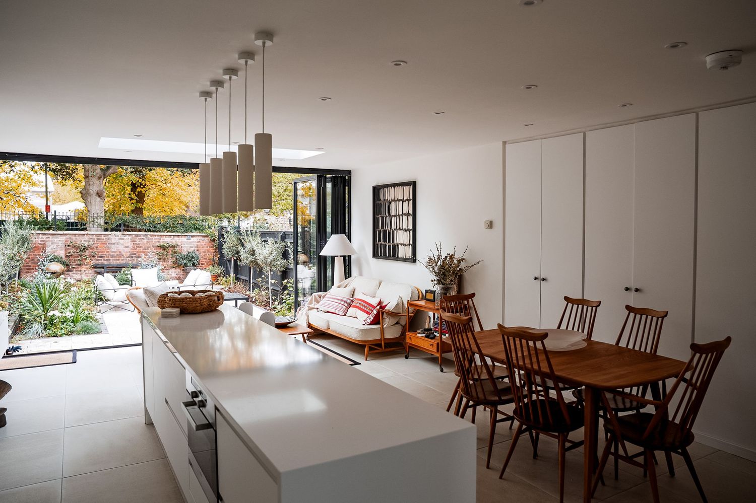 Modern open-plan kitchen and dining area with white cabinetry, wooden furniture, and large glass doors opening to a lush garden.