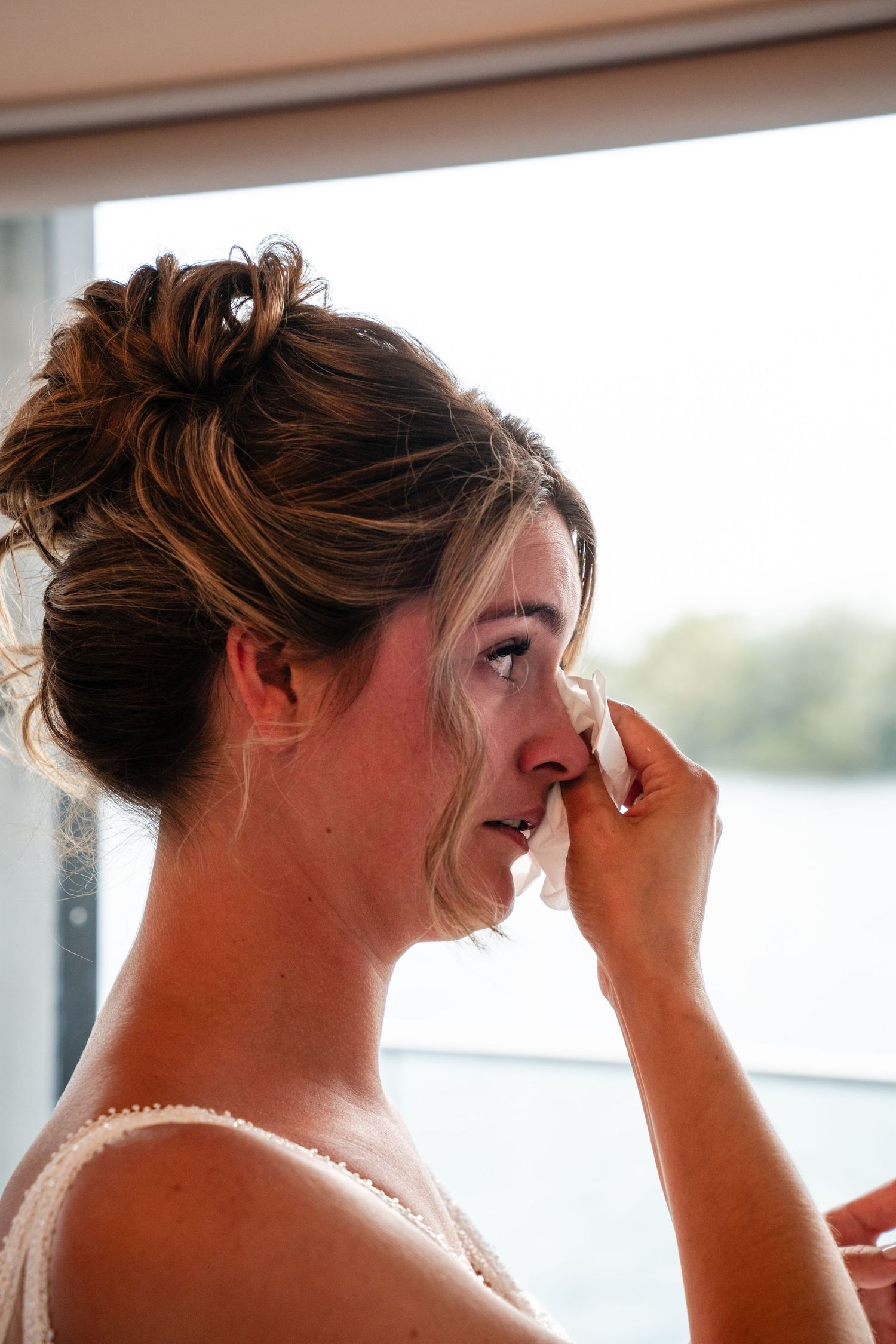 Emotional bride wiping tears with tissue, elegant updo hairstyle, seated near window with soft natural light.