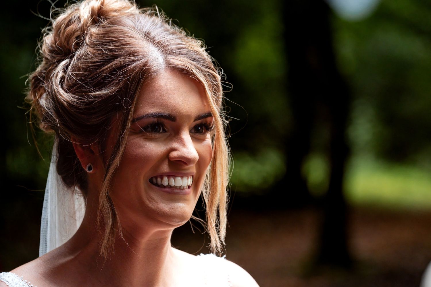 Smiling bride with elegant updo hairstyle and veil, captured outdoors with soft natural light on her wedding day.