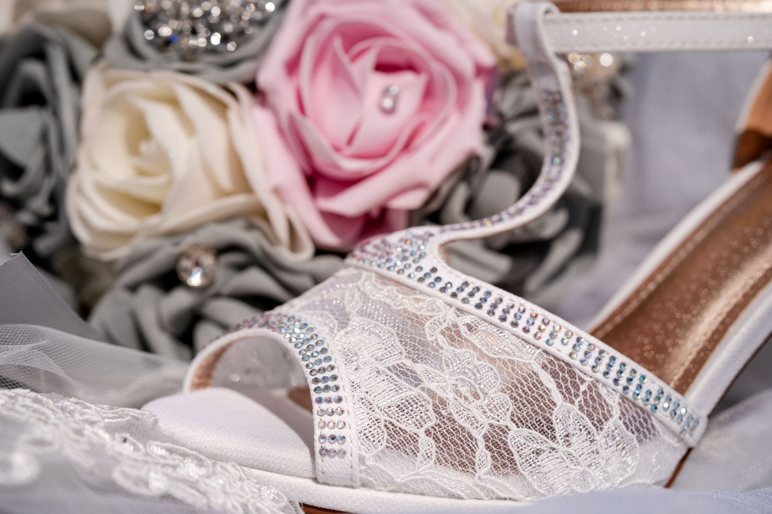 White lace bridal heels with rhinestone trim displayed beside pink and cream roses for a wedding.