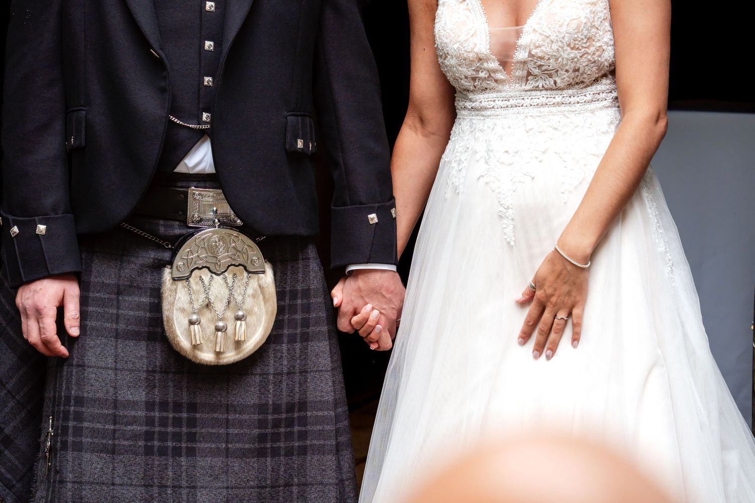 Bride and groom holding hands, groom wearing traditional Scottish kilt with sporran, bride in white lace wedding gown.