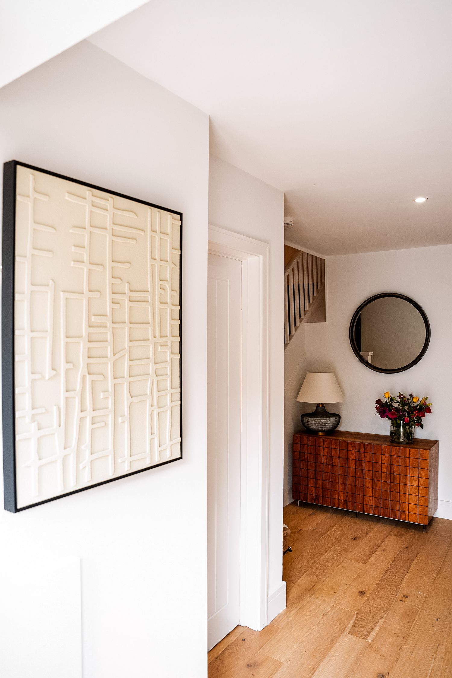 Modern hallway with textured wall art, wooden sideboard, round mirror, and fresh flowers on light hardwood floors.