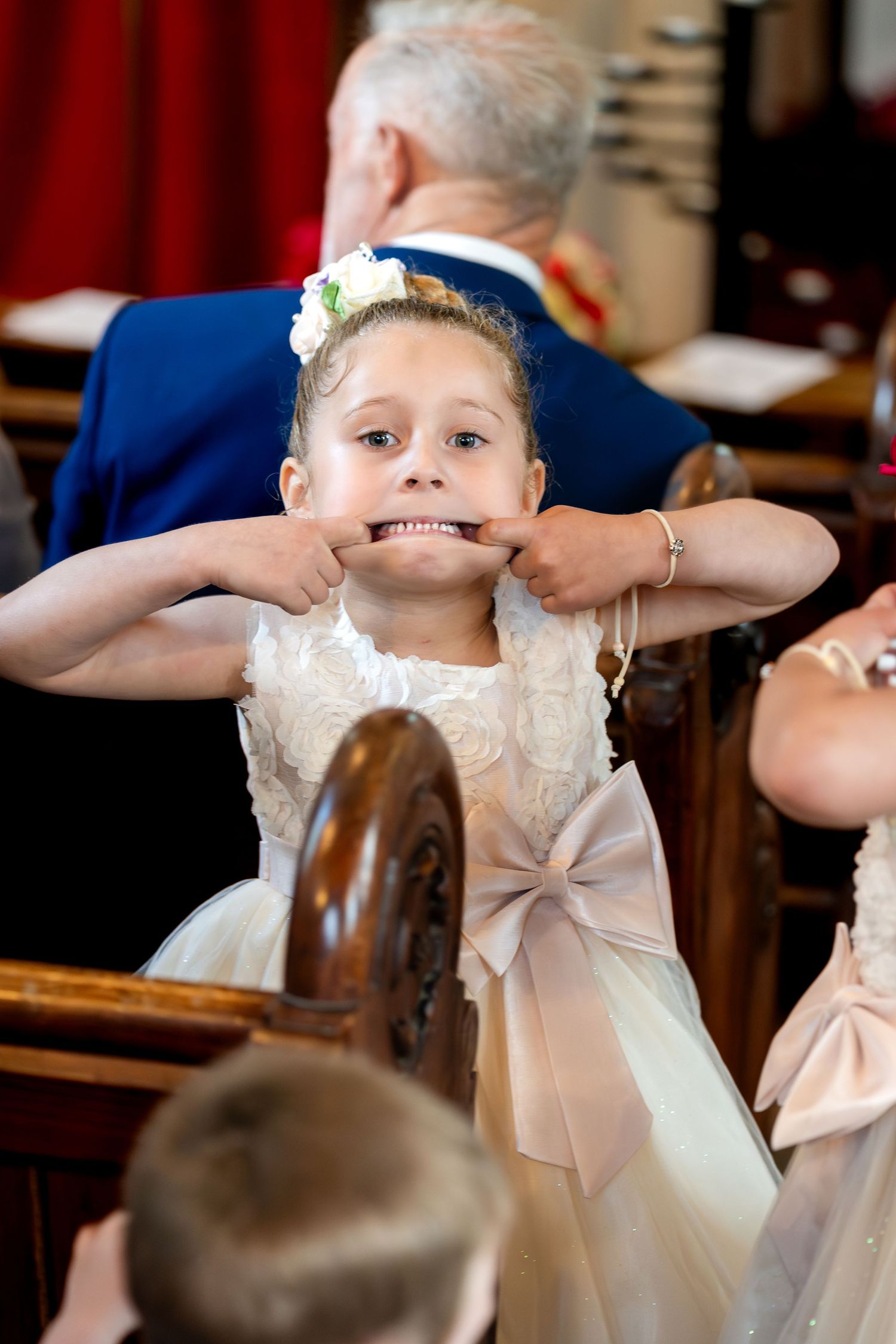 Young flower girl in white dress making funny face during wedding ceremony, pulling cheeks with fingers.