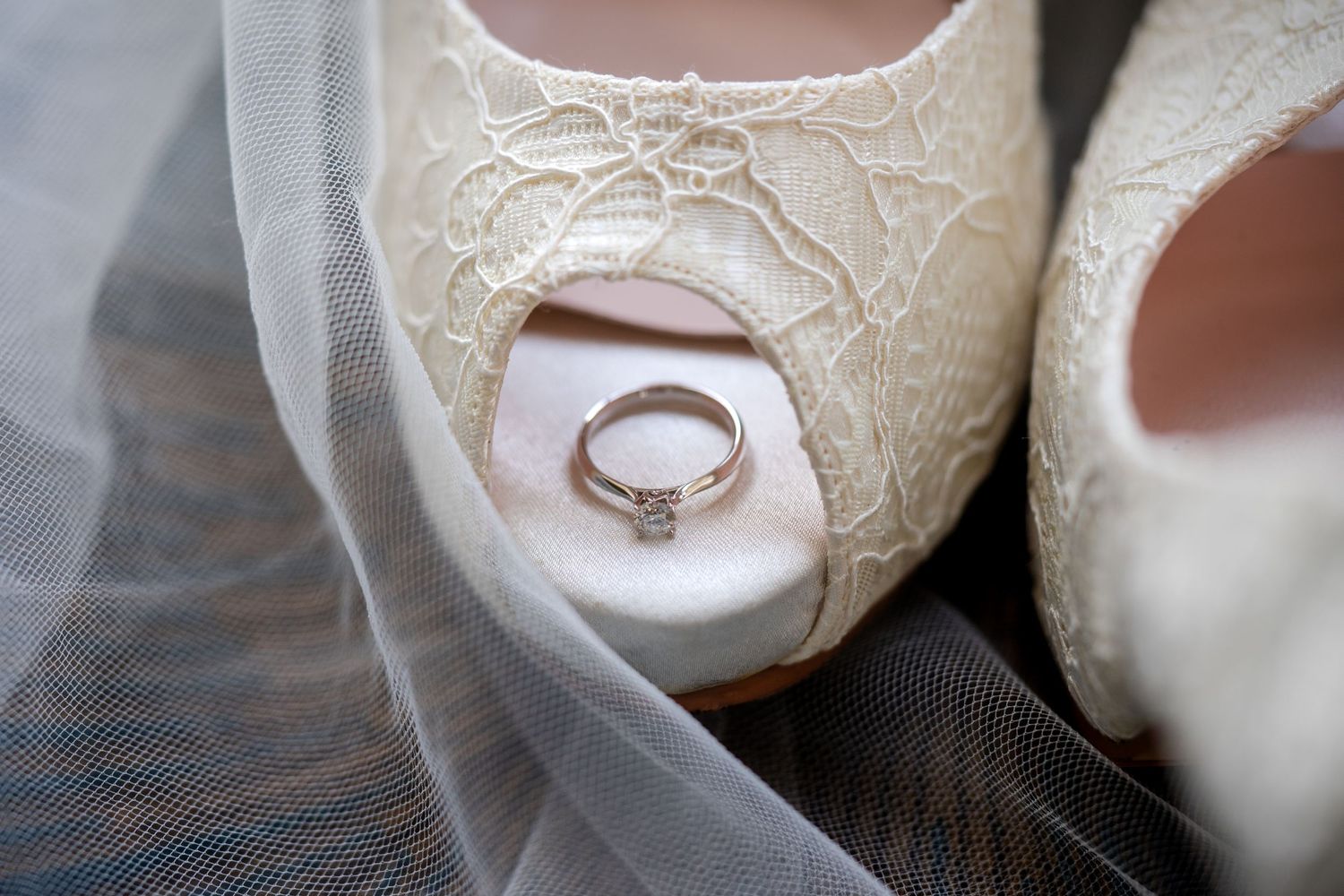 White lace bridal shoes with a diamond engagement ring resting between them on a soft wedding veil.