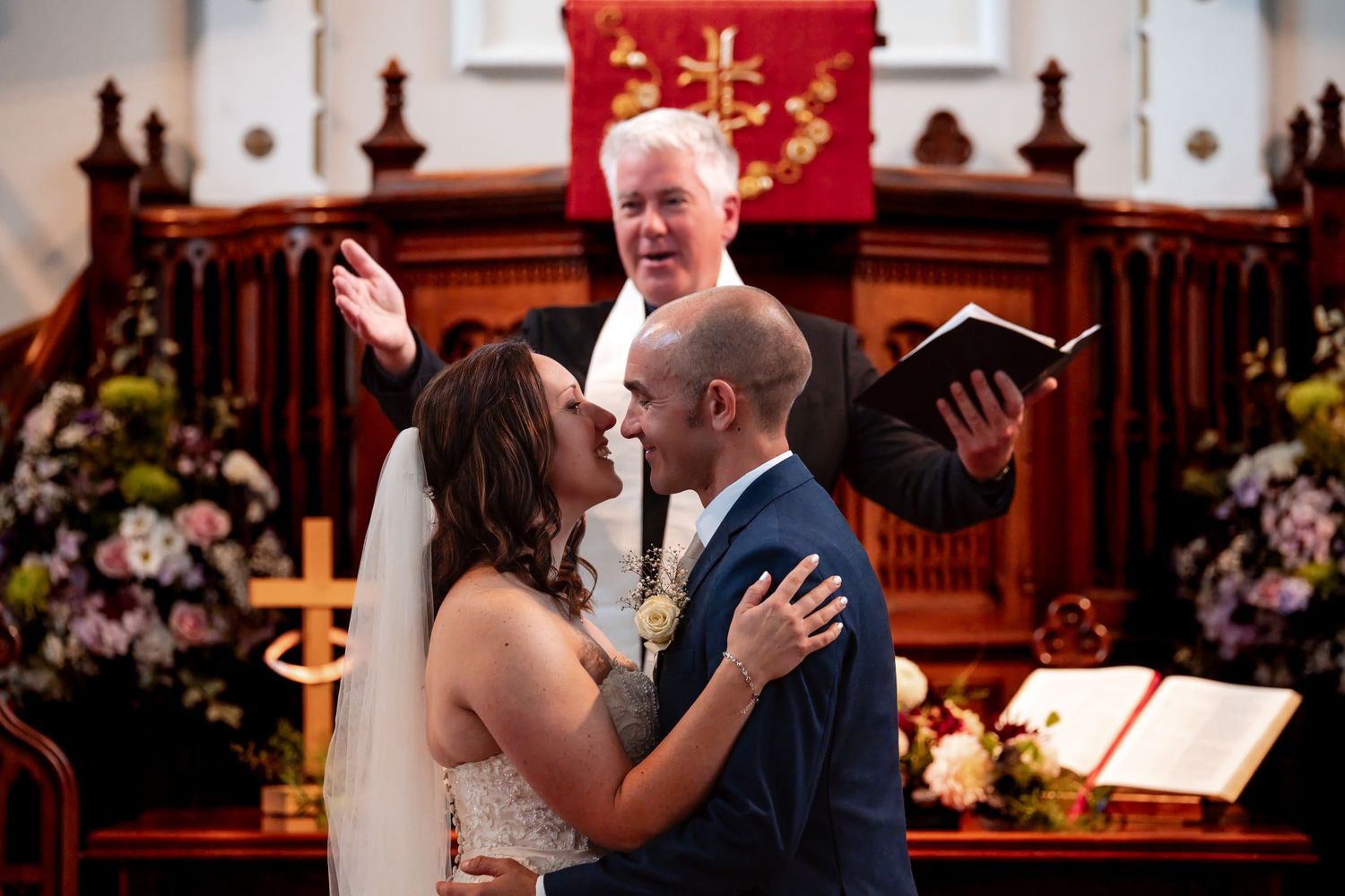 Bride and groom share first kiss at church altar while officiant raises hand in blessing during wedding ceremony.