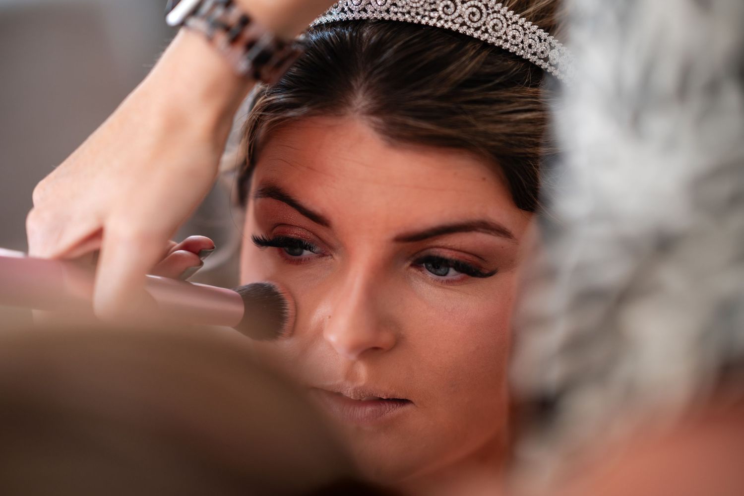 Bride wearing a tiara gets professional makeup applied with a brush before her wedding ceremony.