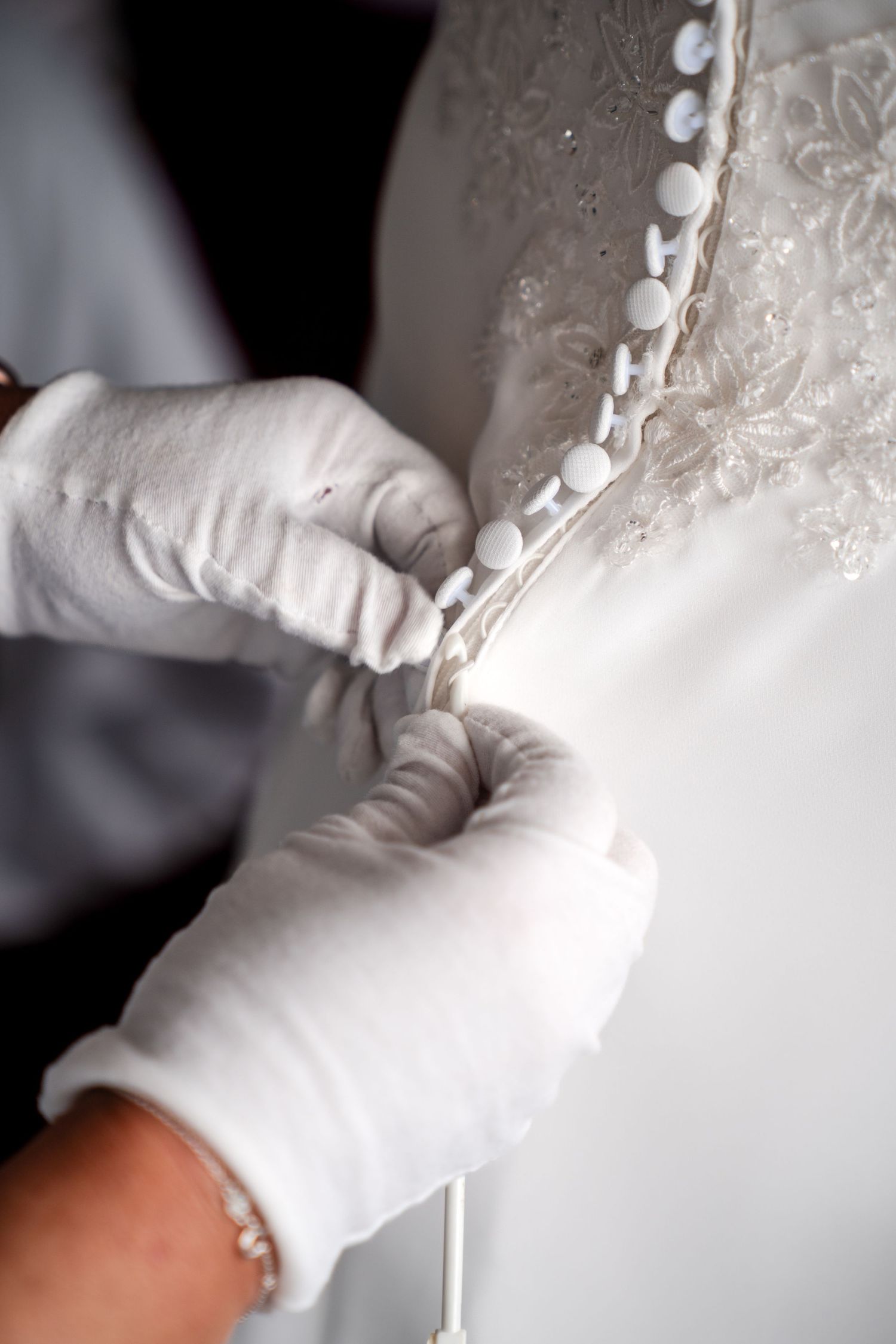 Gloved hands carefully fastening small buttons on the back of an elegant white lace wedding dress.