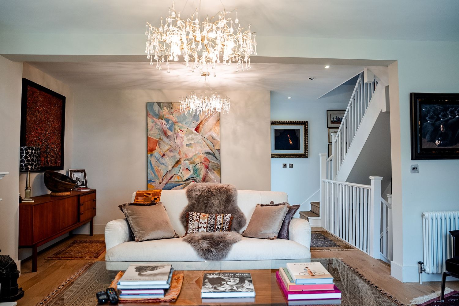 Elegant living room with crystal chandelier, white sofa, colorful abstract art, and staircase in a stylish home interior.