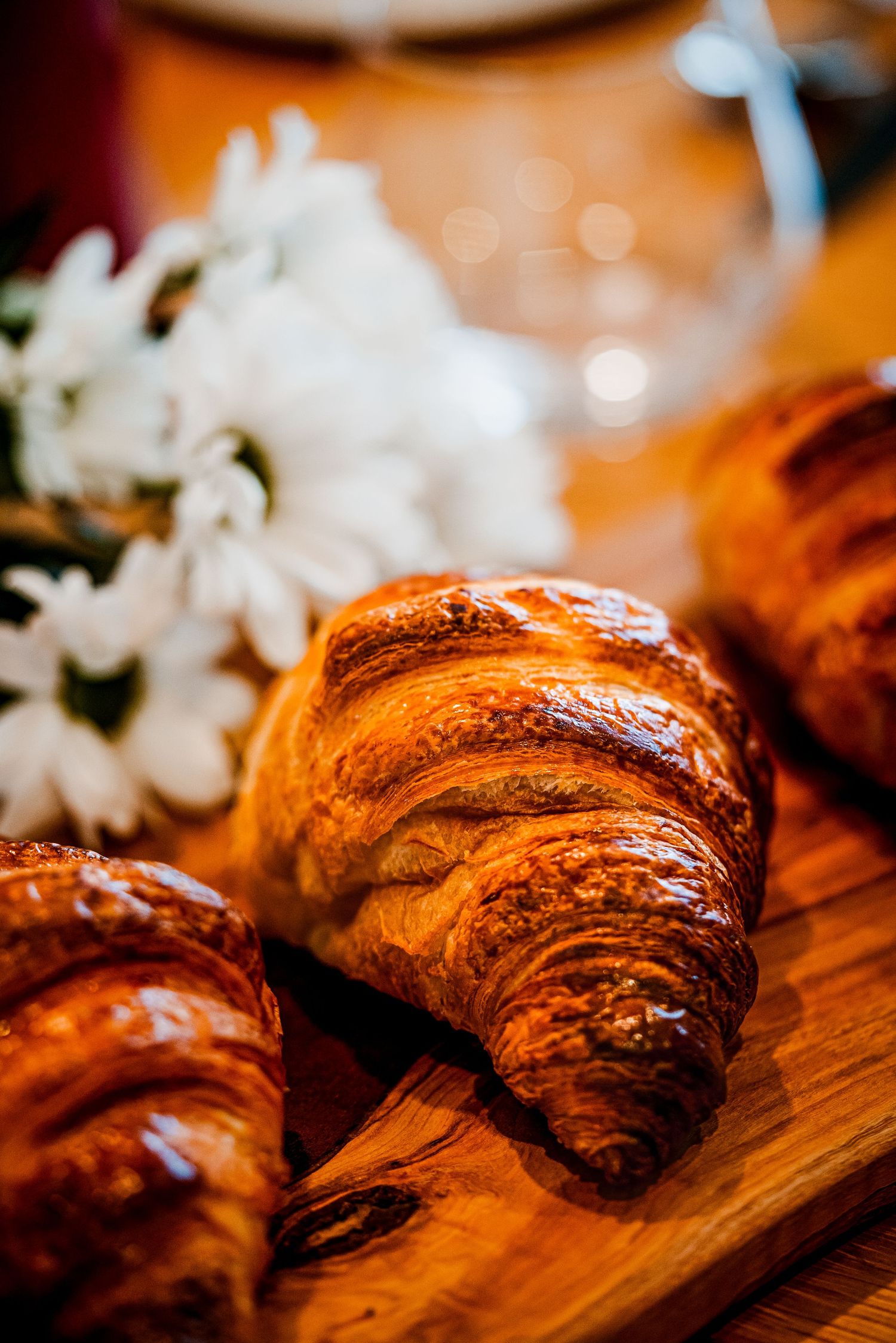Golden croissants on a wooden board with white flowers in soft bokeh background.