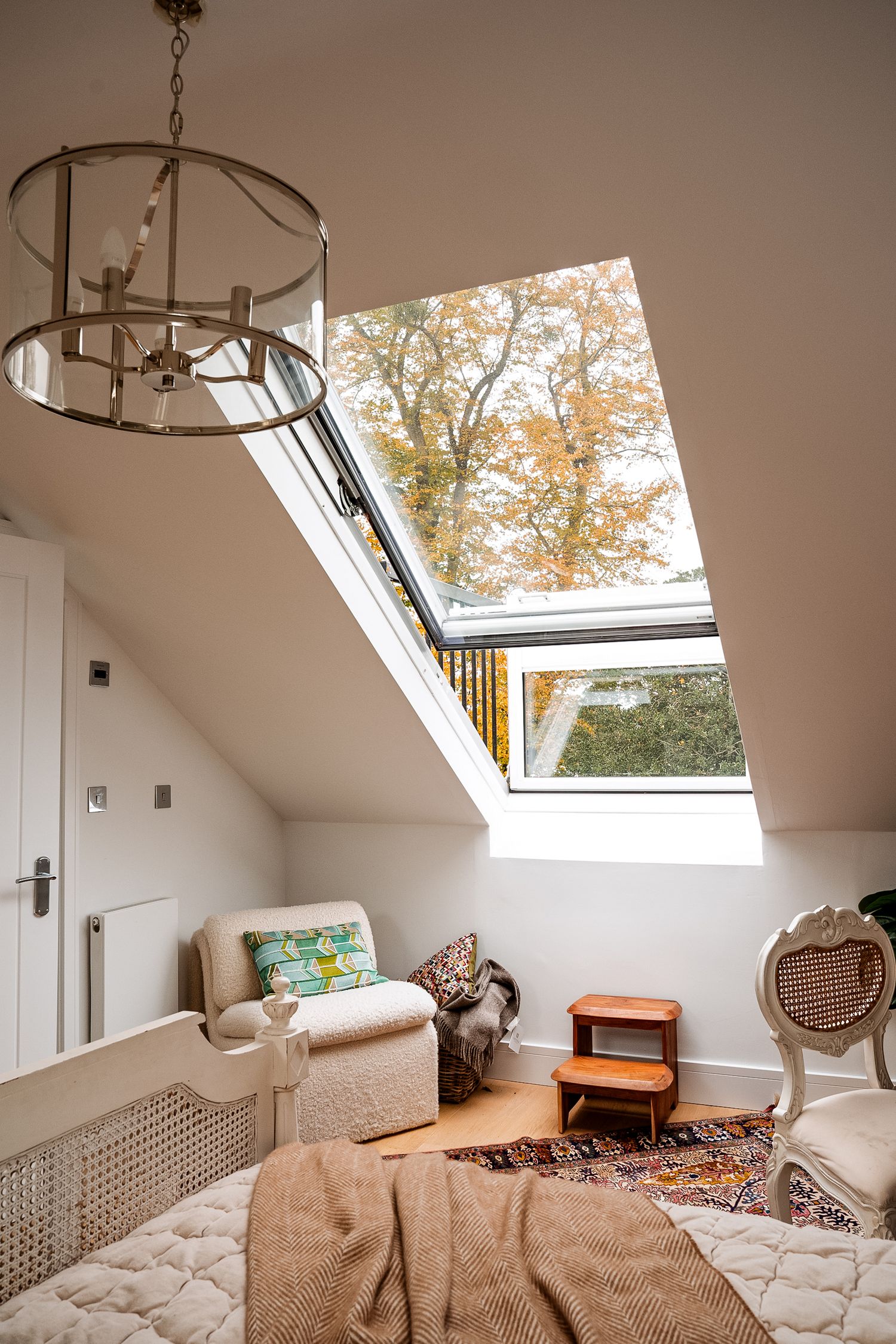 Bright attic bedroom with skylight window showing autumn tree, chandelier, armchair, and cozy decor on hardwood floors.
