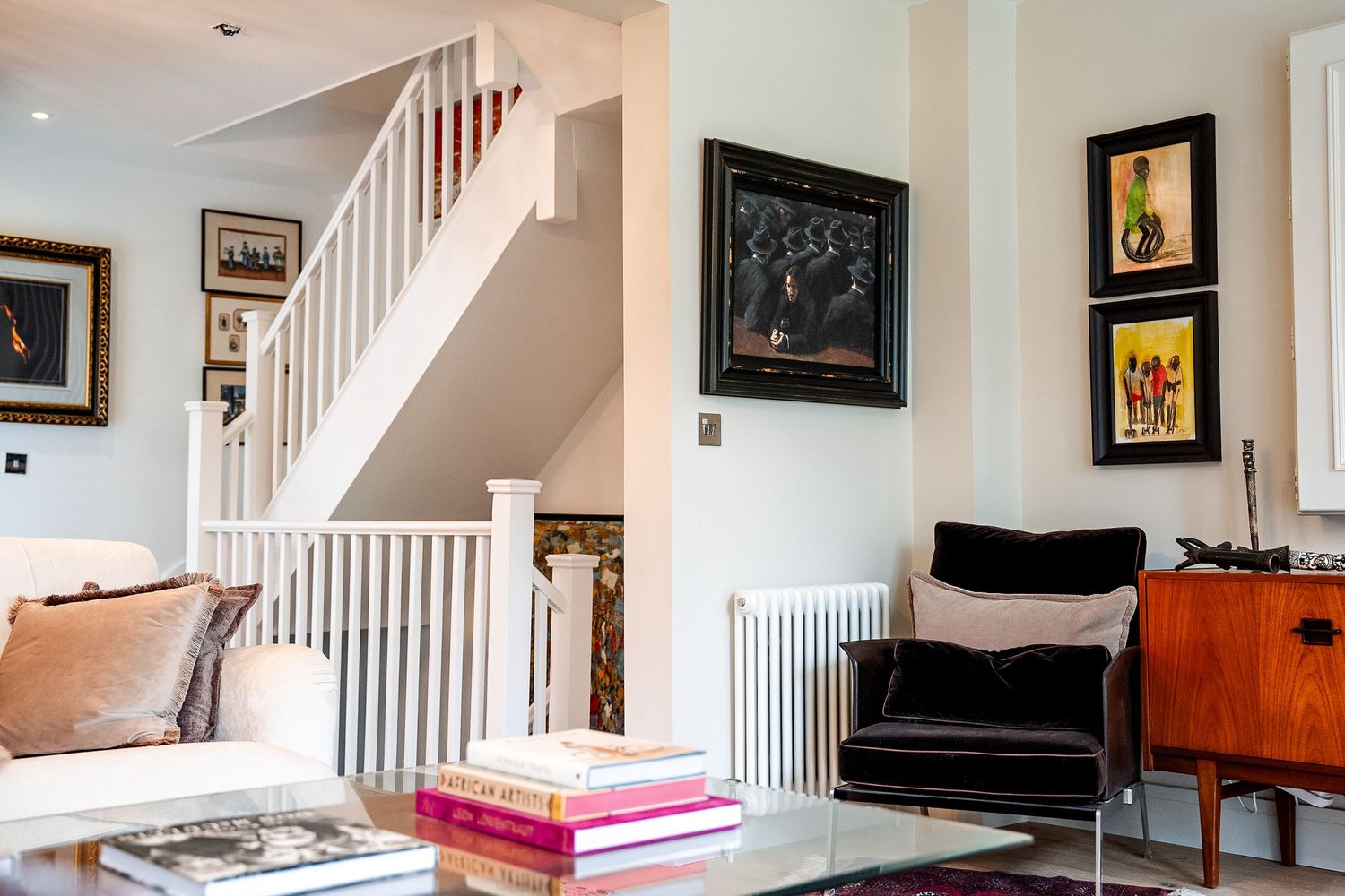 Elegant living room with white staircase, black velvet armchair, glass coffee table, and colorful artwork on white walls.