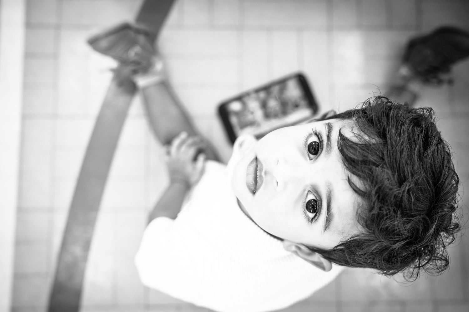 Black and white photo of a young curly-haired child looking up at camera with wide eyes indoors.