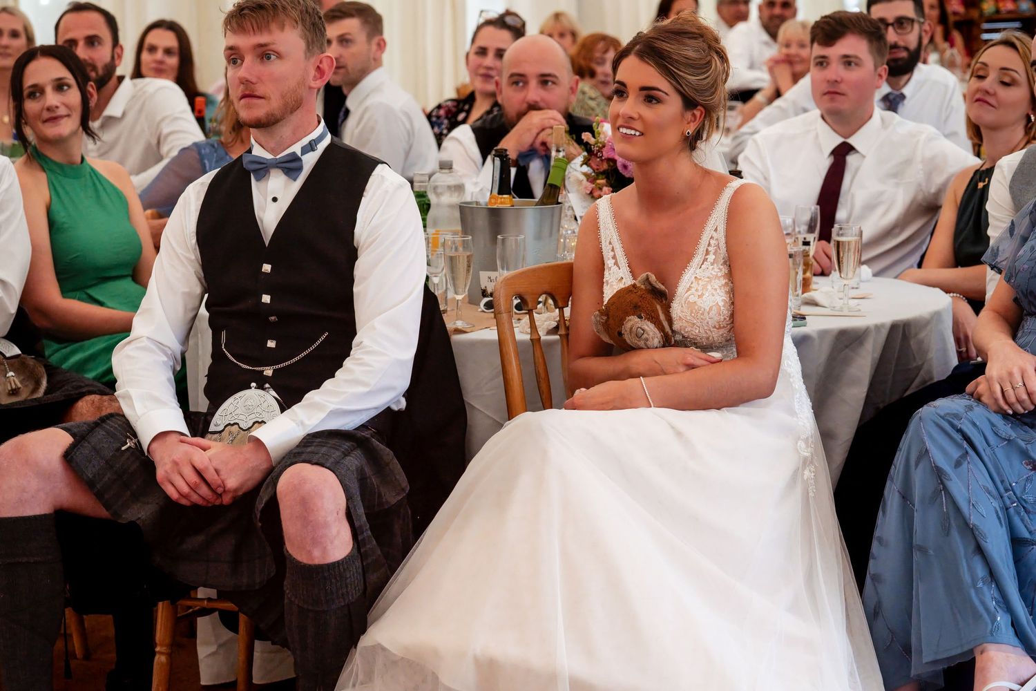 Bride in white gown and groom in Scottish kilt sit together at wedding reception surrounded by guests.