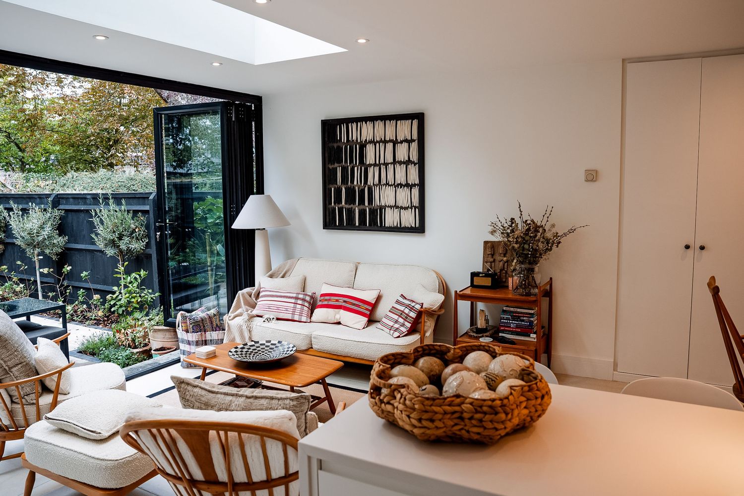 Modern living room with white sofa, wooden furniture, wicker basket, and large glass doors opening to a lush garden.