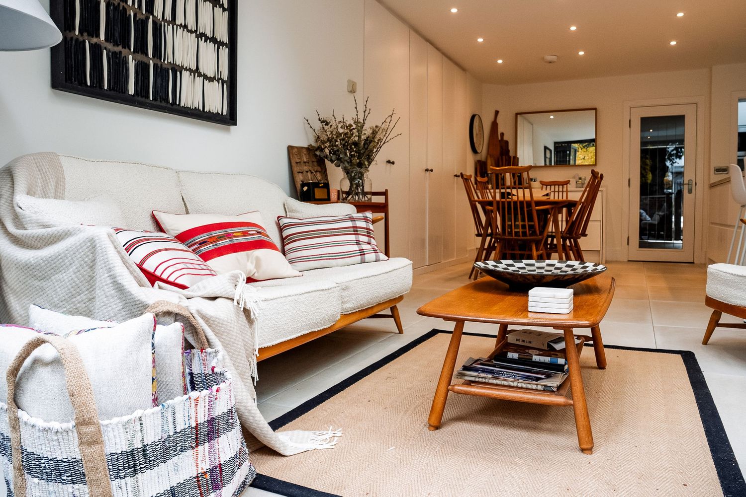 Bright living room with white sofa, red cushions, wooden coffee table, books, and dining area in background.