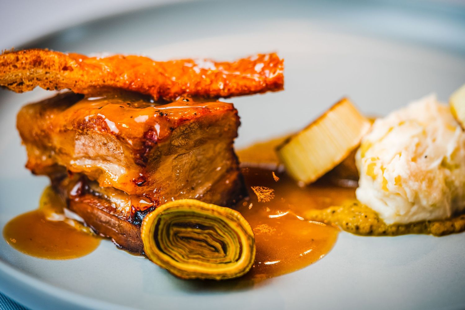 Gourmet braised pork belly with rich caramel sauce, artichoke, crispy crackling, and creamy mashed potato on a white plate.