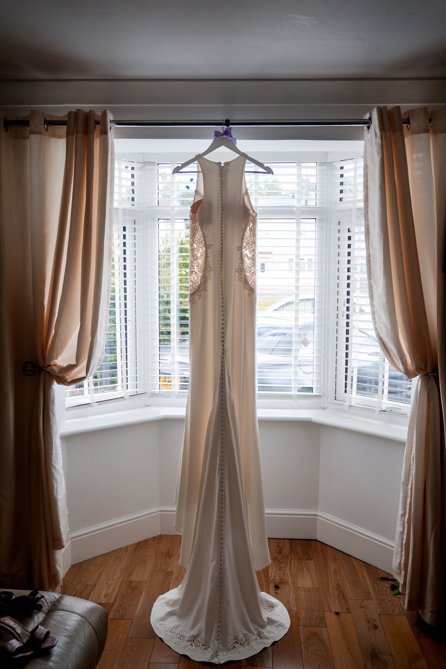 Elegant white bridal gown with lace back detail hanging in a bay window with wooden floors and neutral curtains.