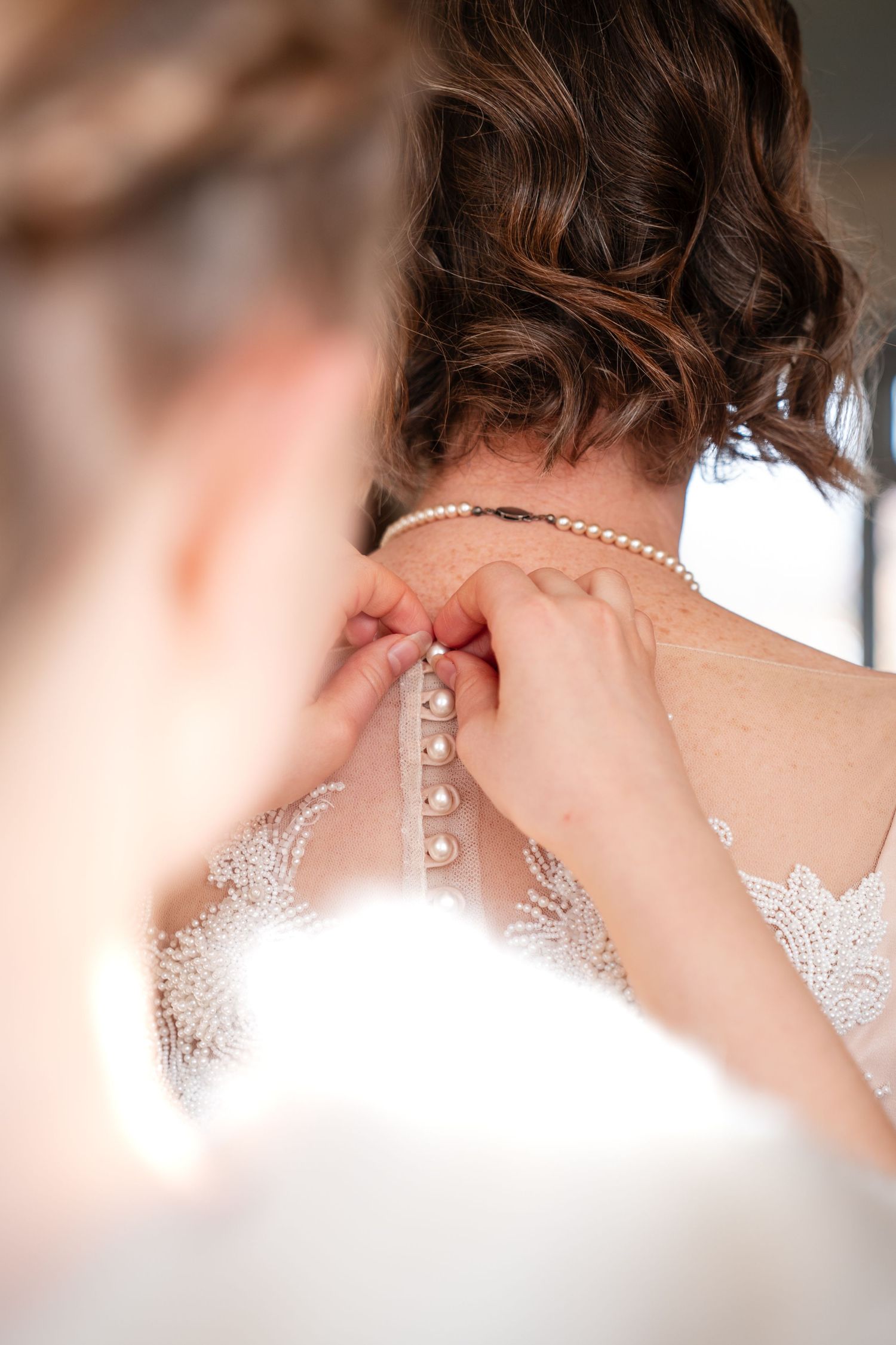 Bridesmaid fastening pearl necklace on bride in lace wedding dress, close-up of wedding preparation moment.