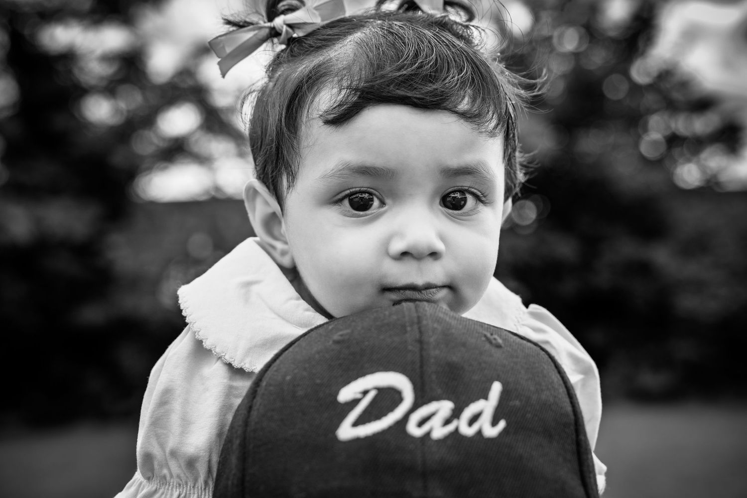 Black and white photo of a cute toddler girl with a bow holding a baseball cap that reads Dad.
