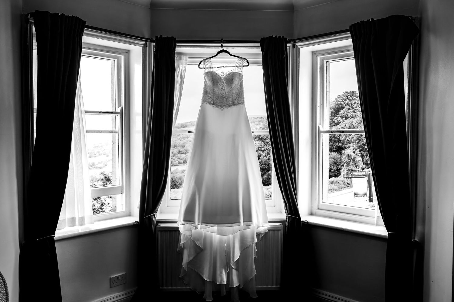 Black and white photo of an elegant wedding dress hanging in a bay window with dark curtains.