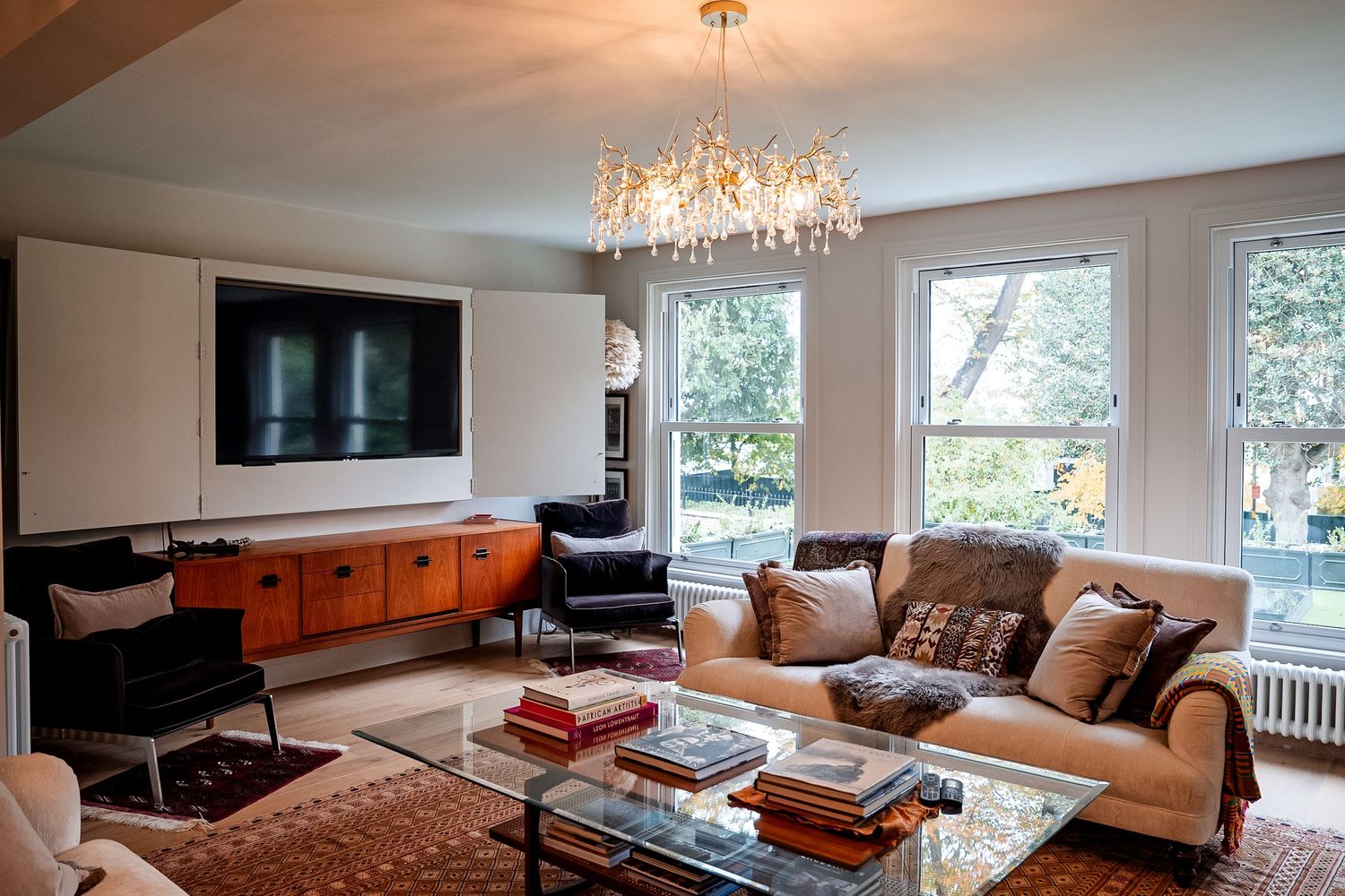 Elegant living room with beige sofa, glass coffee table, wood credenza, wall-mounted TV, and gold chandelier near bay windows.