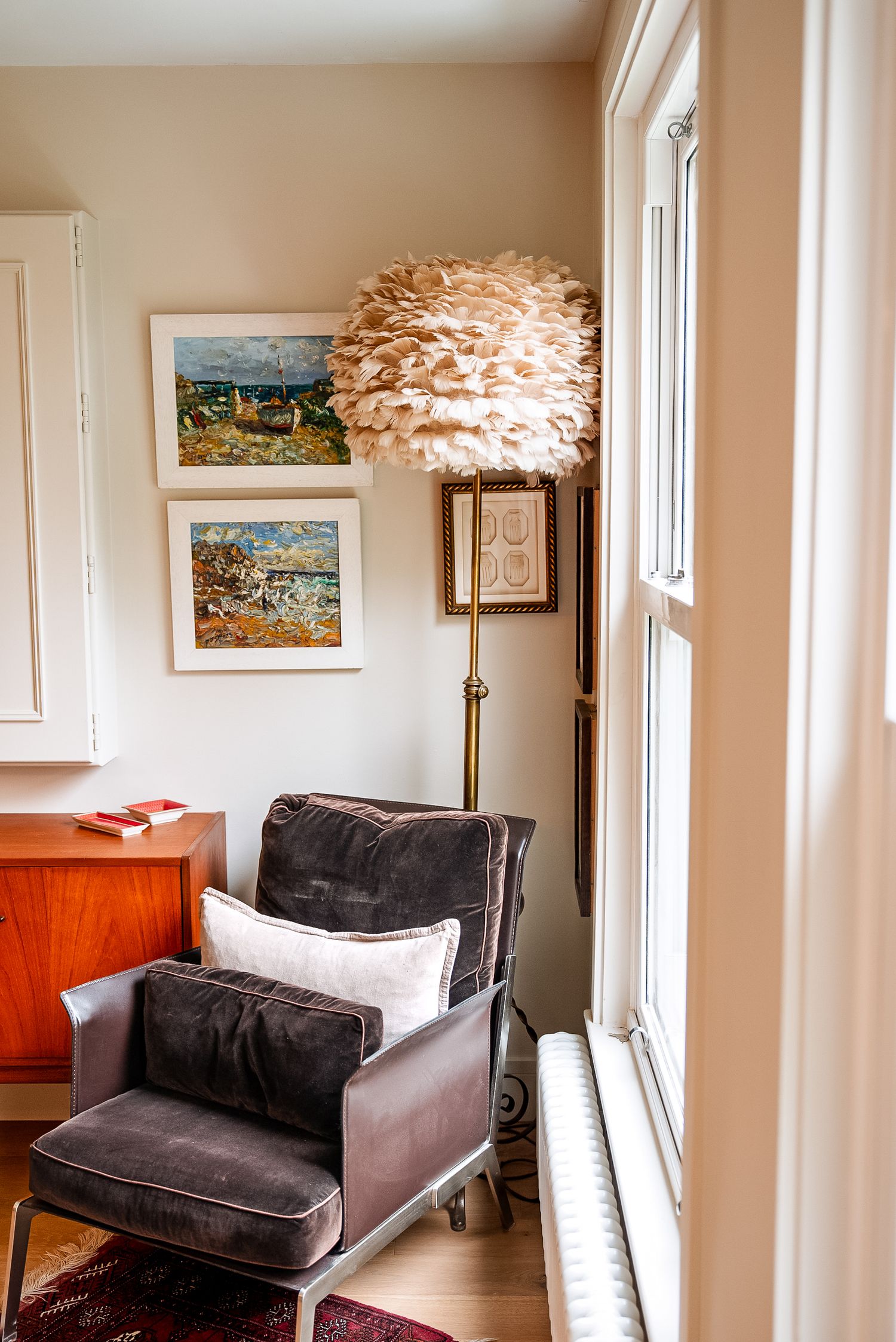 Cozy reading nook with gray velvet armchair, feather floor lamp, and colorful framed artwork on beige walls.