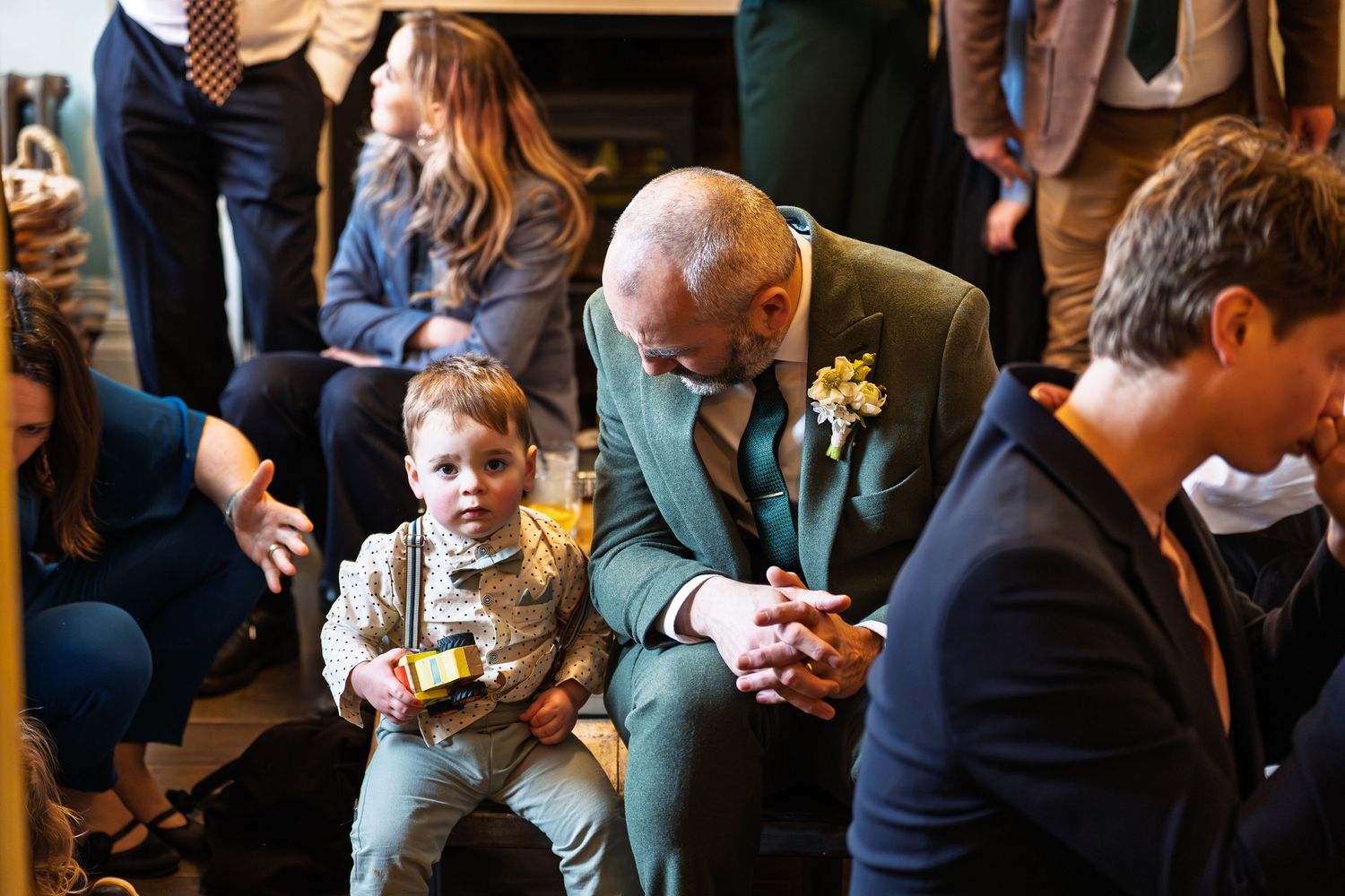 A bald groom in a teal suit chats with a young boy in suspenders at a wedding reception.