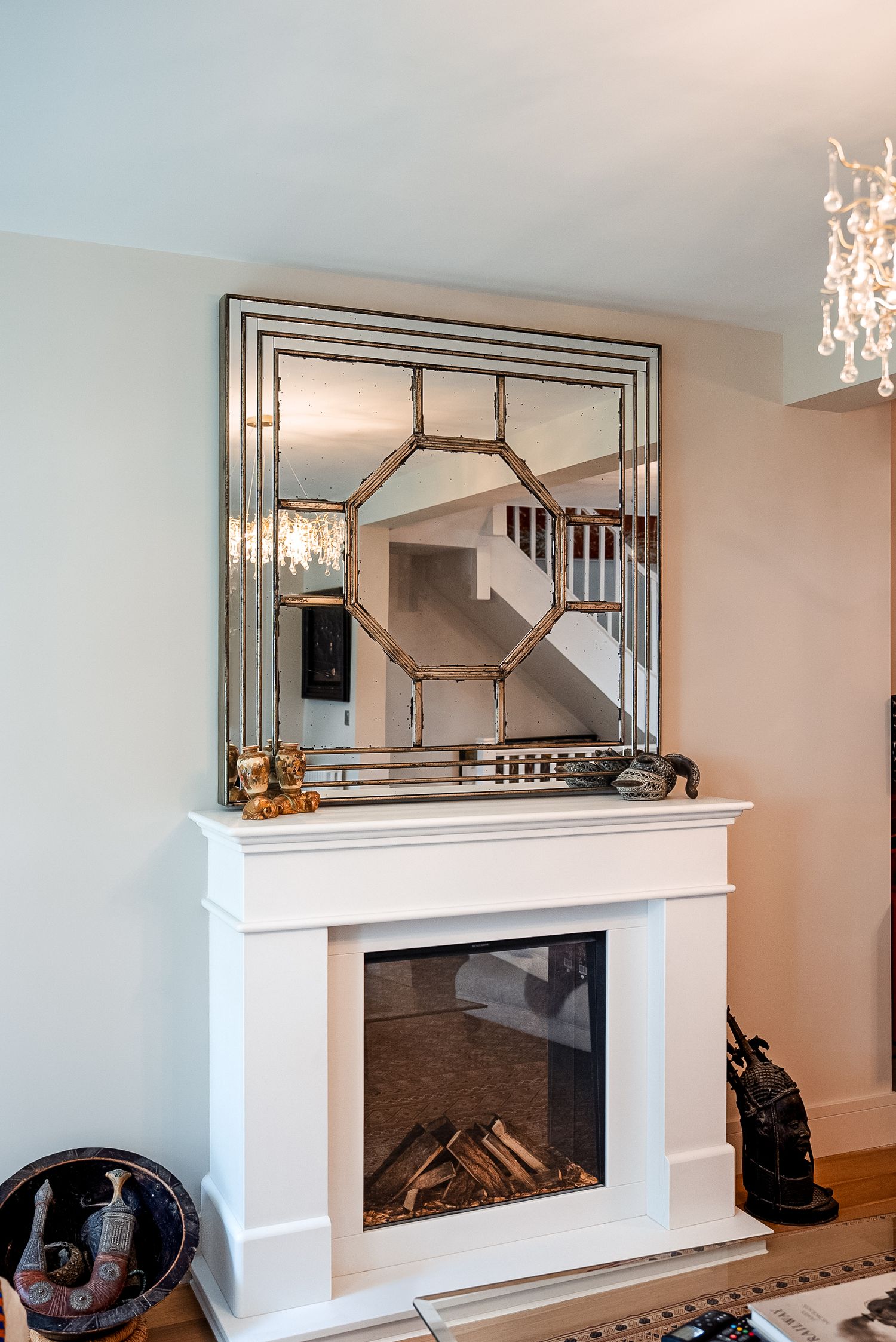 White fireplace with decorative mirror above mantel, wood logs inside, and elegant chandelier visible in background.