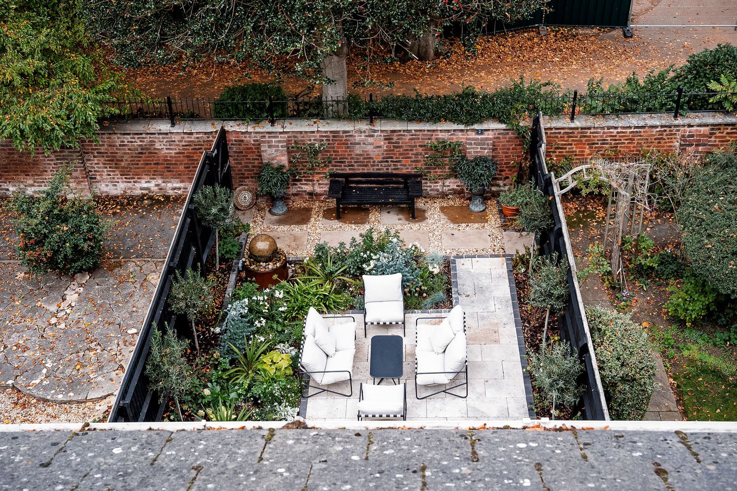 Aerial view of a walled garden with white lounge chairs, lush greenery, and a decorative fountain on a stone patio.