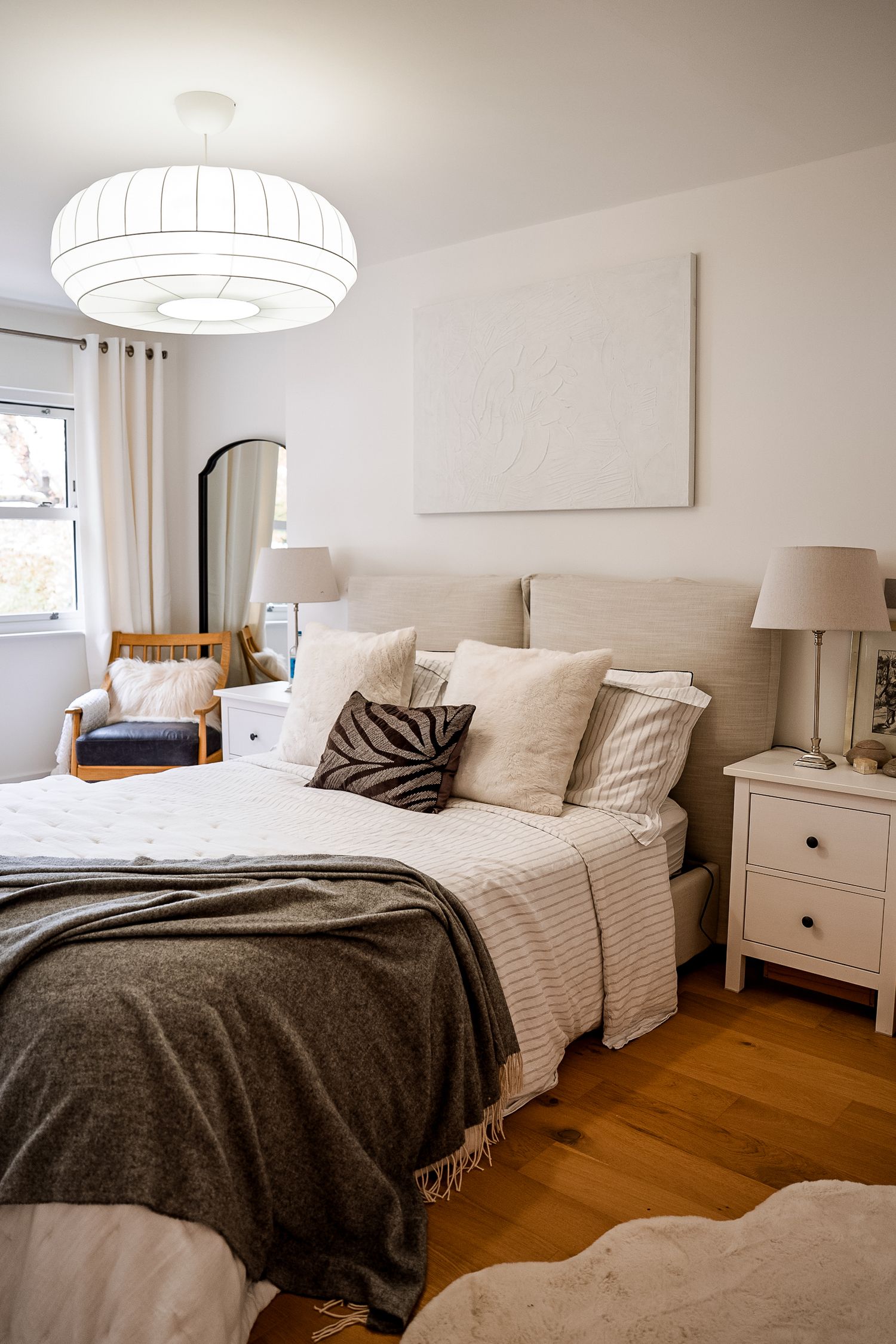 Cozy neutral bedroom with beige upholstered bed, layered pillows, gray throw blanket, white nightstands, and warm hardwood floors.