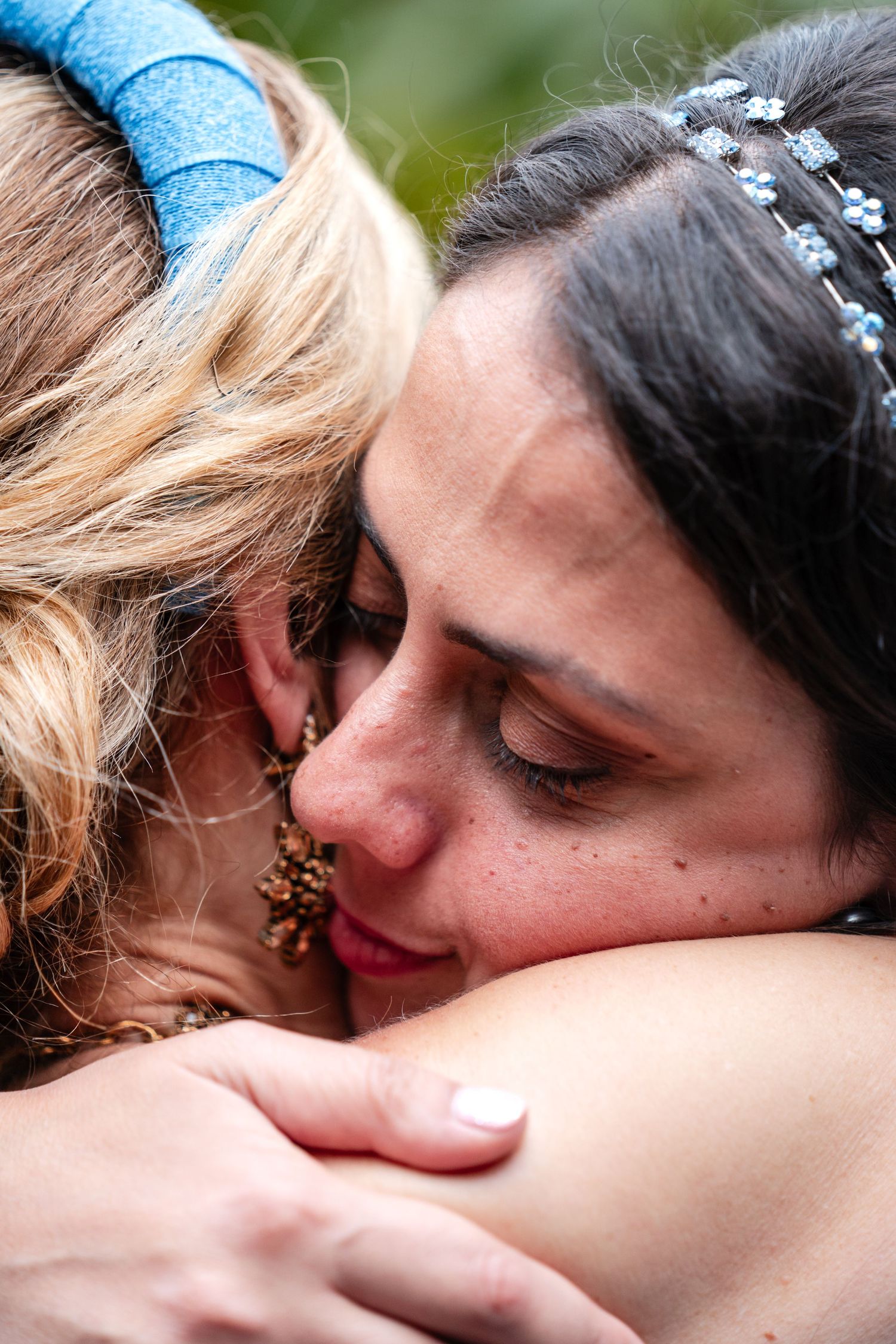 Two women sharing an emotional hug, one with a blue headband and one with a floral hair accessory, eyes closed.