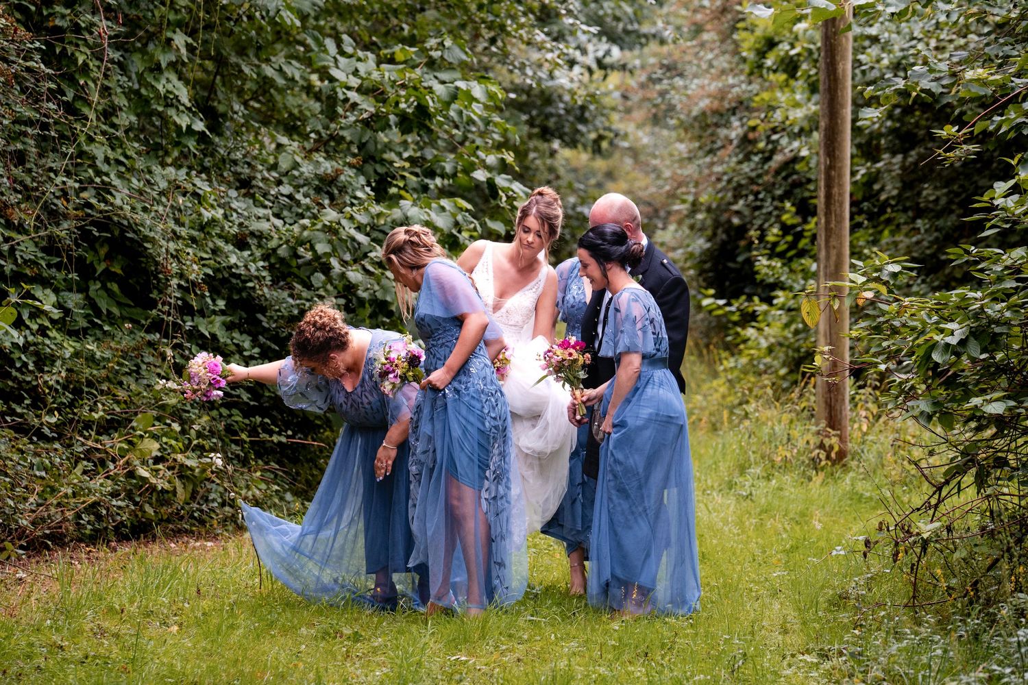 Bride in white gown with bridesmaids in blue dresses walking through lush green garden path at outdoor wedding.