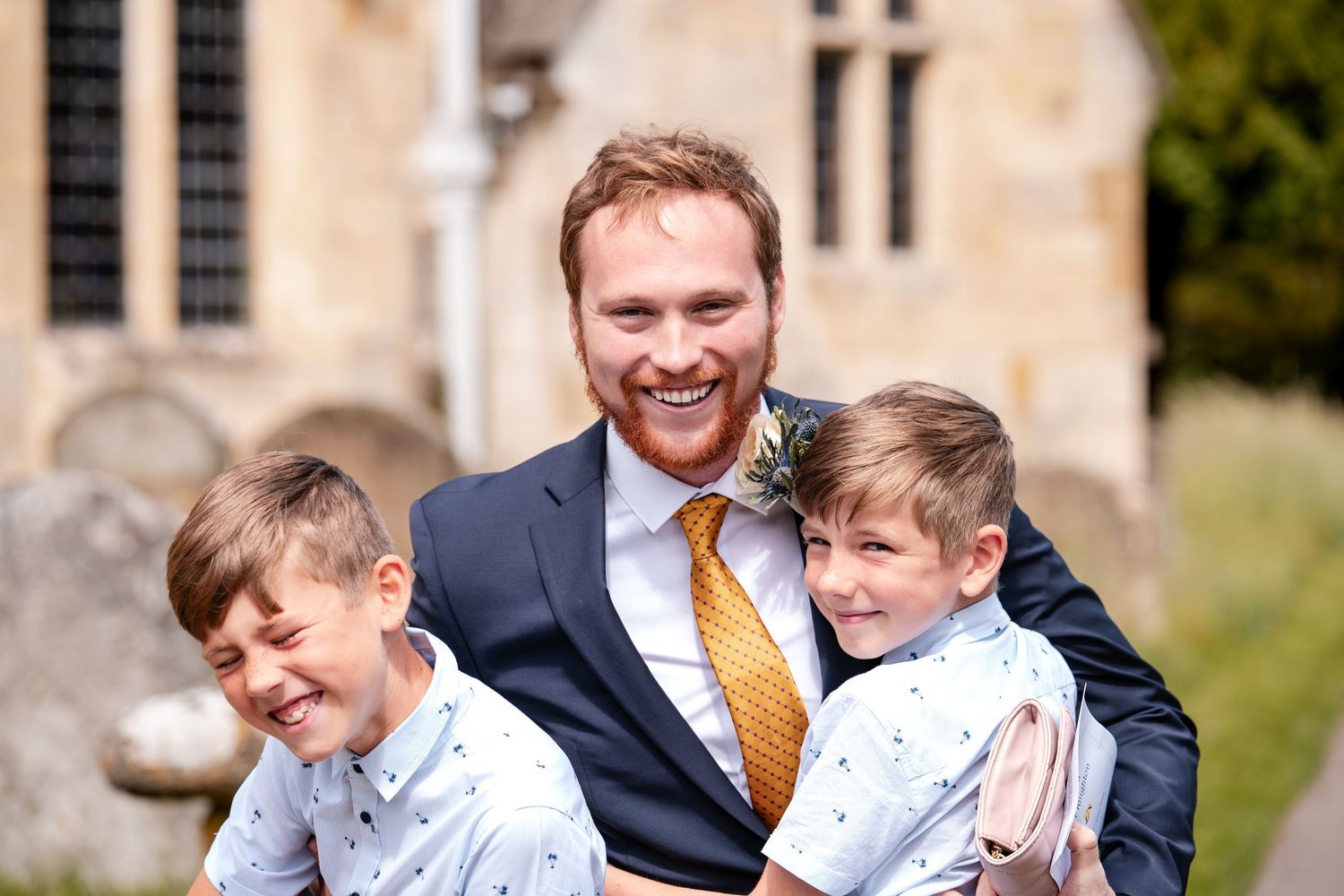 Smiling groom in navy suit and gold tie hugs two young boys in matching shirts outside a church.