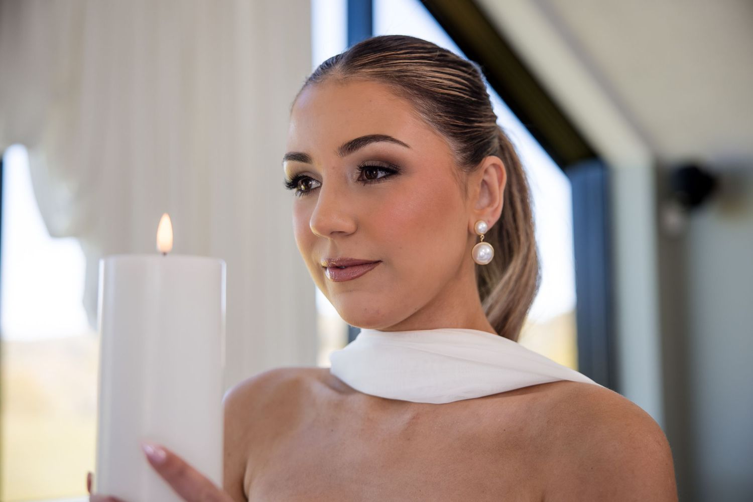 Elegant woman in bridal look holding a white pillar candle, wearing pearl earrings and a white wrap.