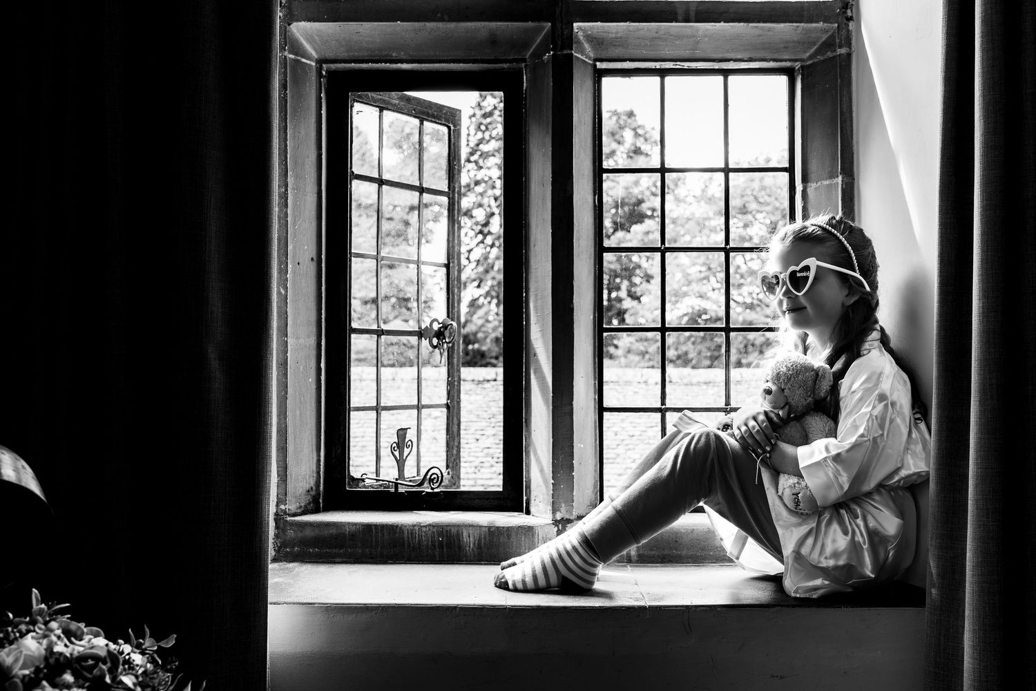 Black and white photo of a child sitting on a windowsill wearing heart-shaped sunglasses, gazing thoughtfully outside.