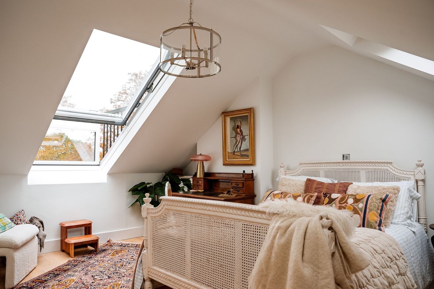 Cozy attic bedroom with skylights, chandelier, wicker bed frame, bohemian rug, and warm neutral decor.