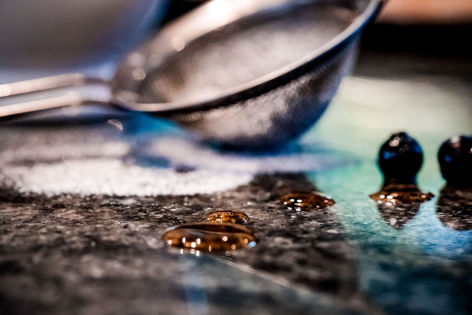 Close-up of a metal spoon with spilled powder, water droplets, and dark berries on a textured surface.