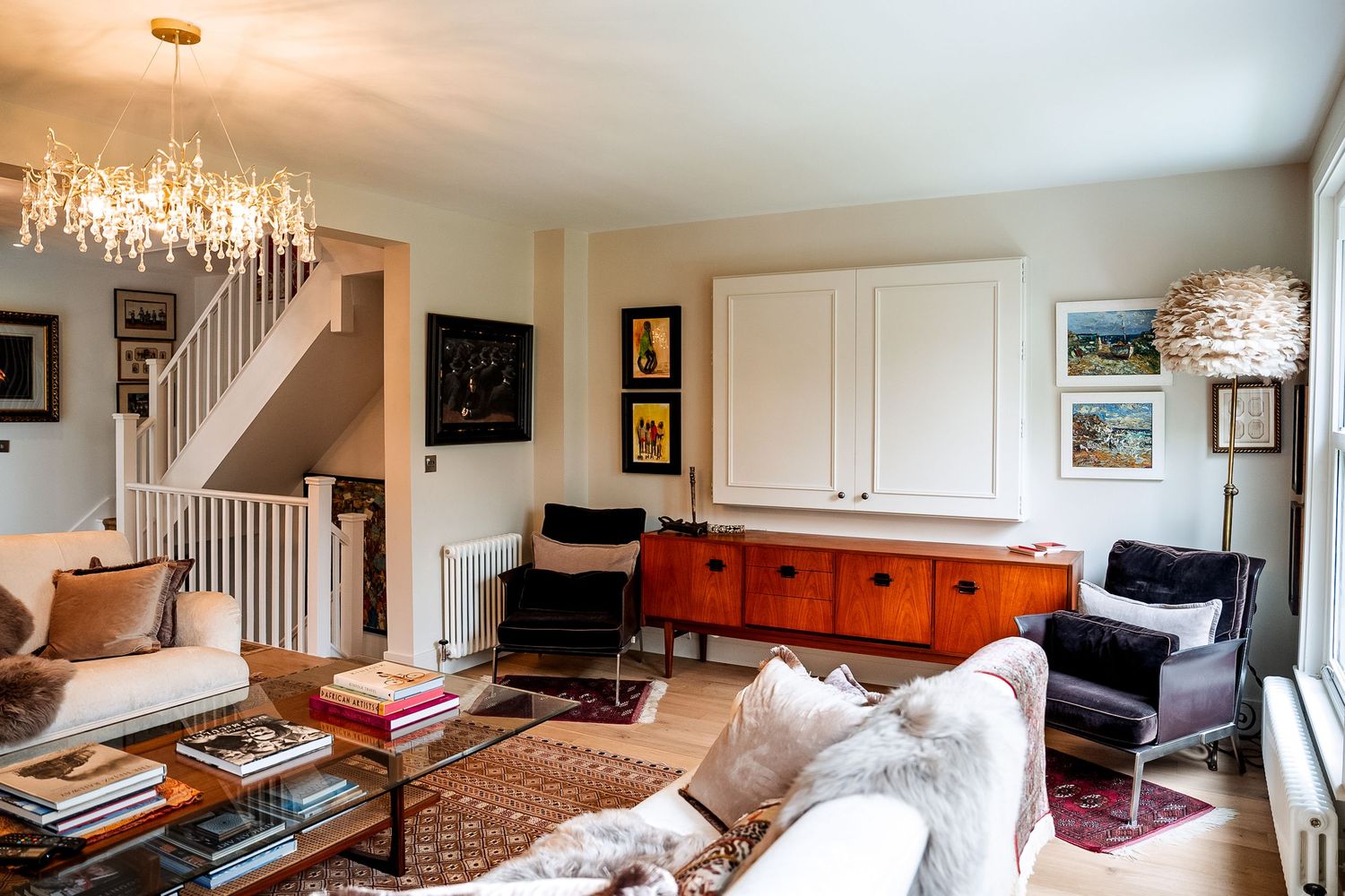 Eclectic living room with chandelier, white sofa, orange sideboard, black chairs, staircase, and colorful artwork on walls.