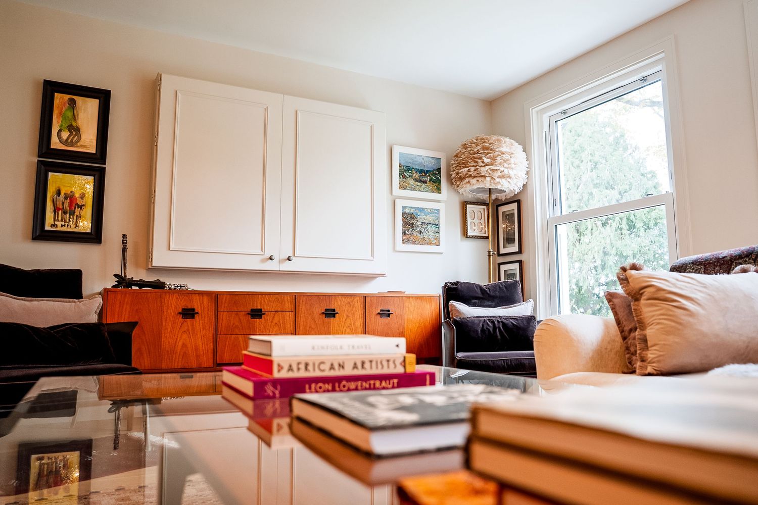 Cozy living room with orange credenza, stacked books, glass coffee table, white cabinets, and a bright window with lush greenery.