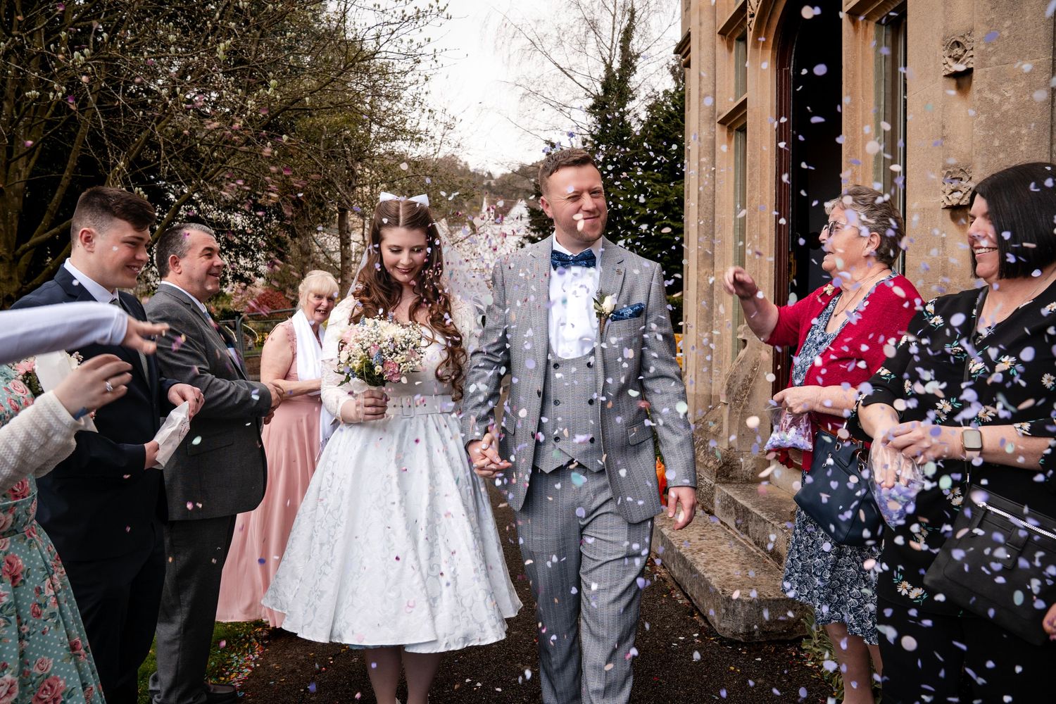 Bride and groom walk through confetti shower surrounded by smiling wedding guests outside a stone building.