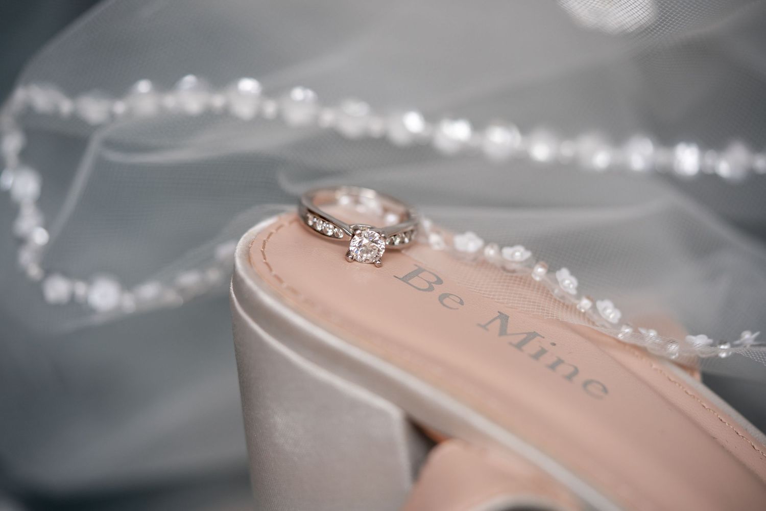 Diamond engagement ring resting on a beige heel inscribed with Be Mine, surrounded by pearl necklace and soft bridal veil.